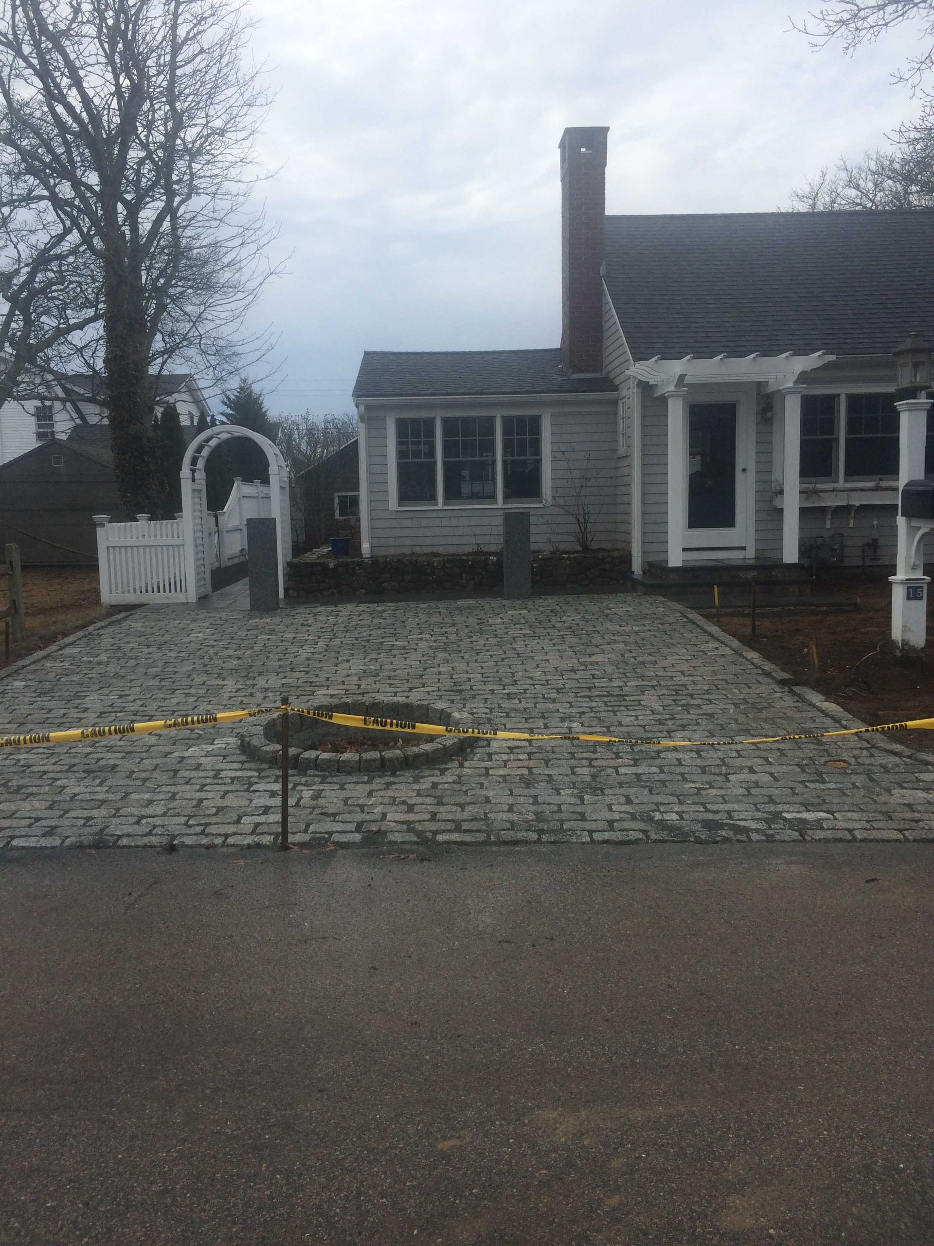 A white house with a cobblestone driveway under construction, featuring a central stone circle and yellow caution tape.
