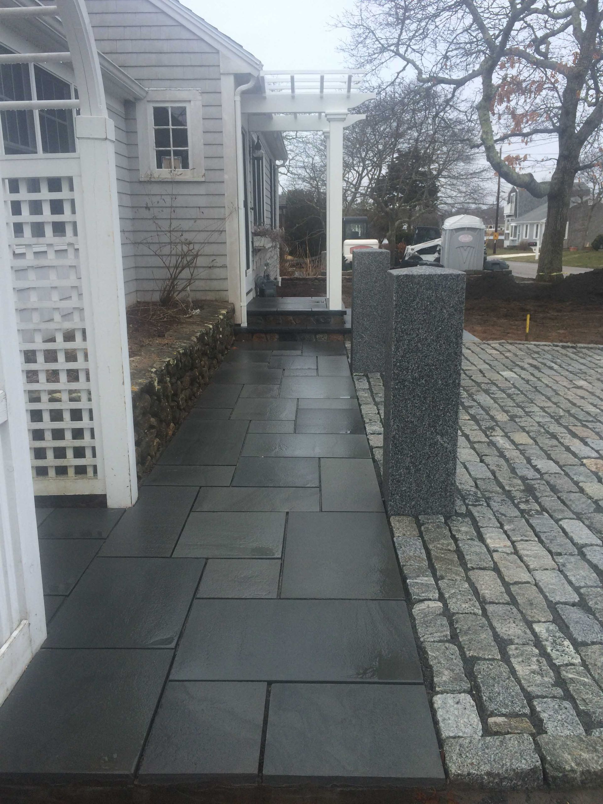 A paved walkway made of dark rectangular stone tiles leads past a light grey house to a white wooden porch entryway.