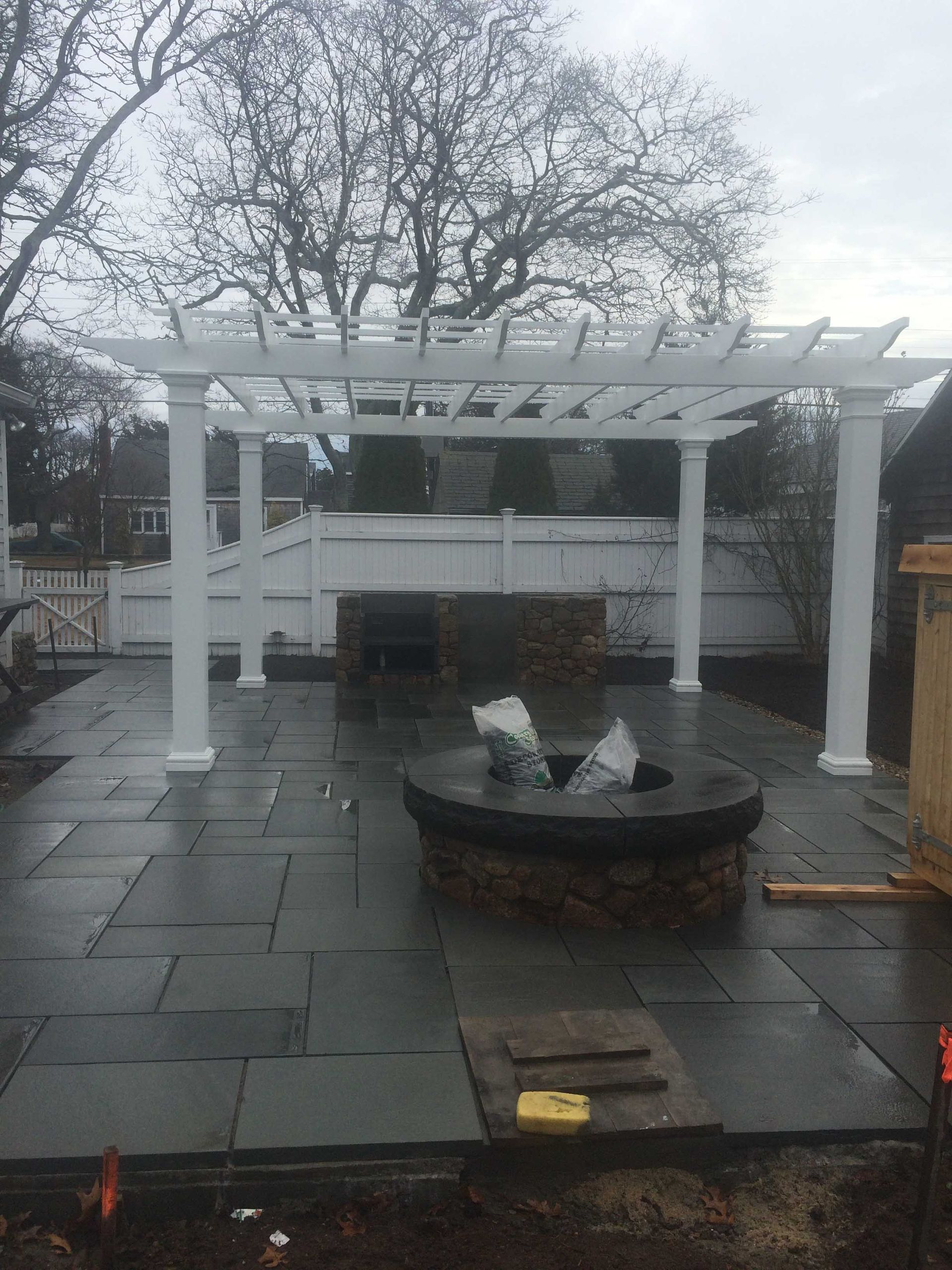 A backyard patio with dark stone pavers, a circular stone fire pit, and a white pergola covering a brick outdoor kitchen.