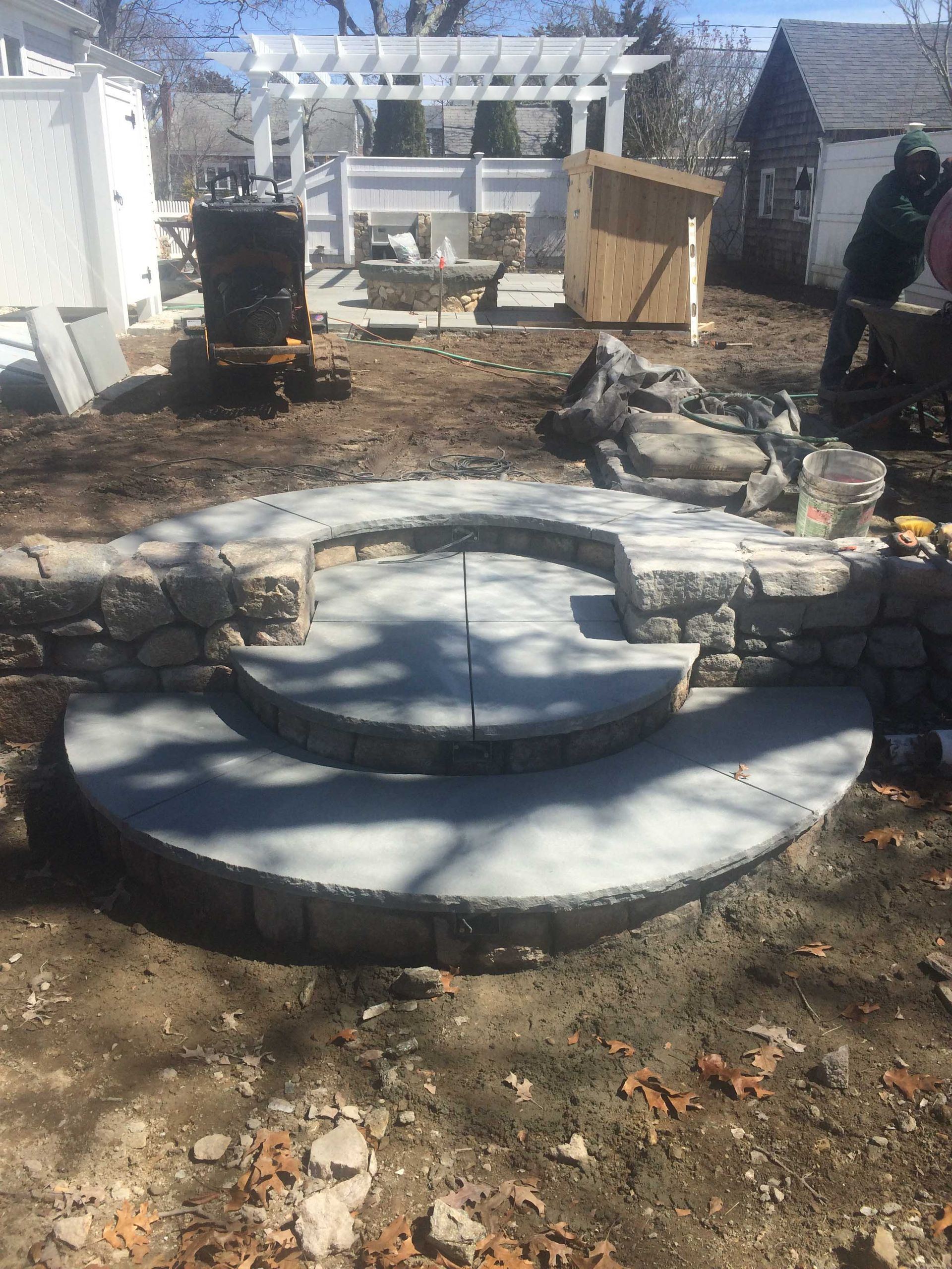 A newly built stone fire pit area with circular steps in a backyard under construction.