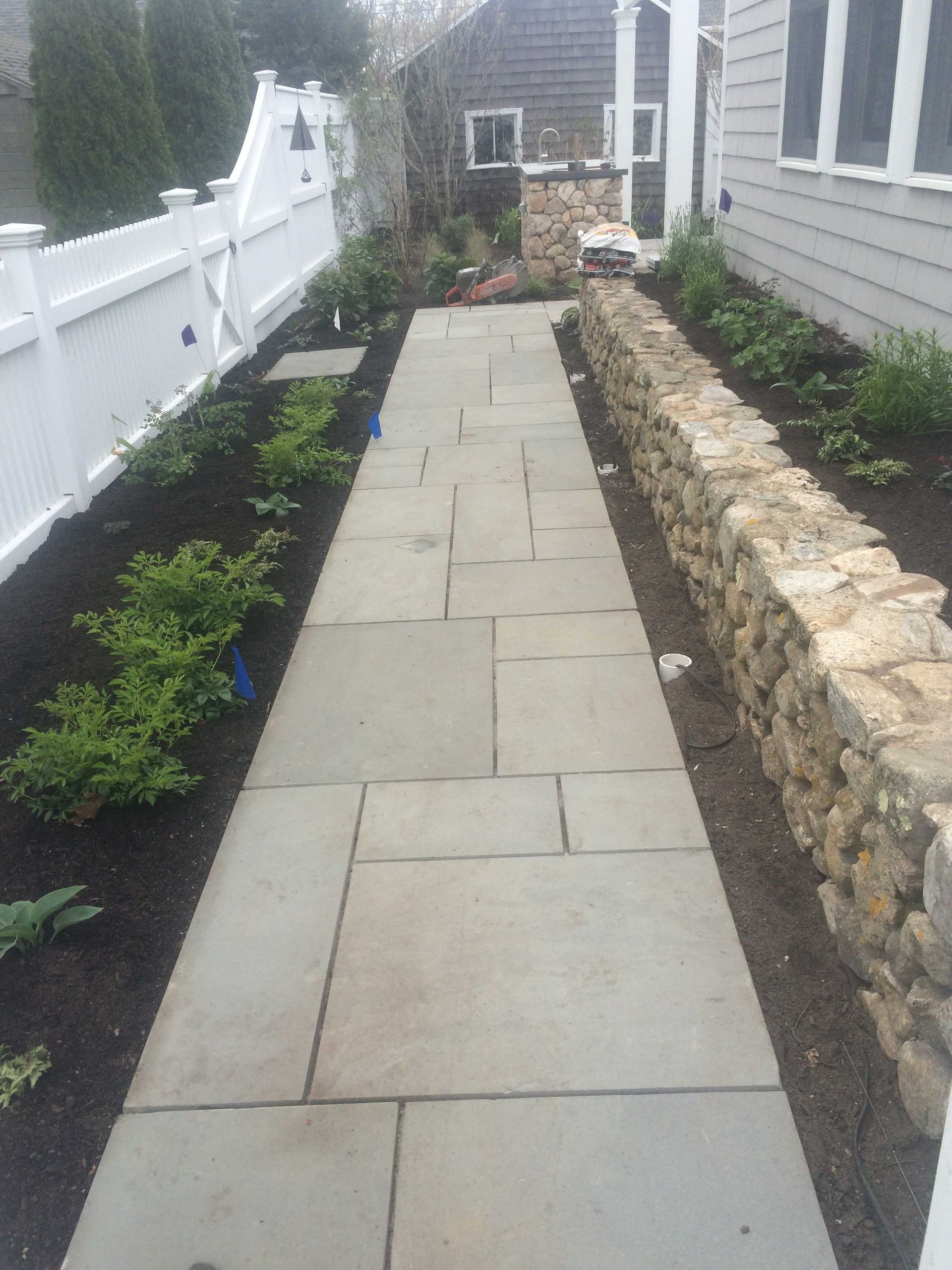 A stone walkway leads toward a house with a white fence on the left and a short stone retaining wall on the right.