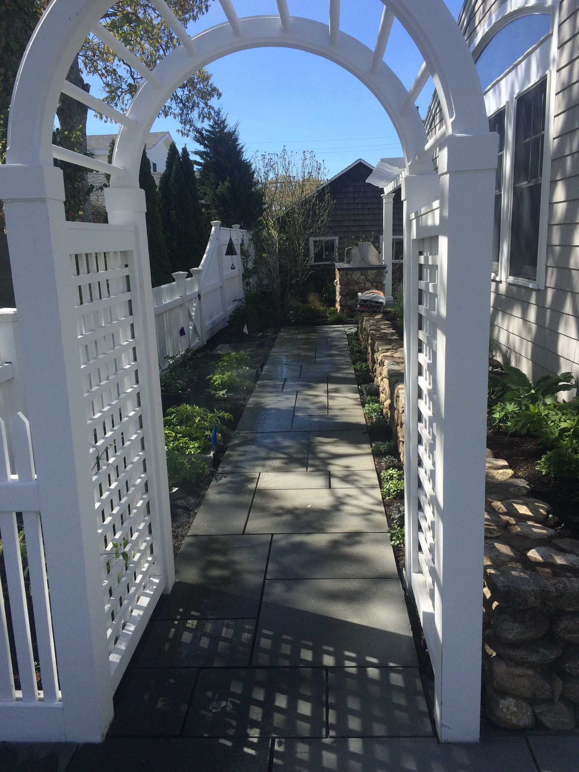 A white wooden garden arbor frames a stone walkway leading to a backyard garden with a white fence and stone landscaping.