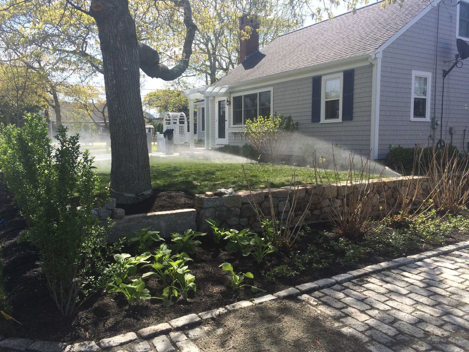 A sprinkler waters a green lawn in front of a gray house with a stone retaining wall and a cobblestone driveway.