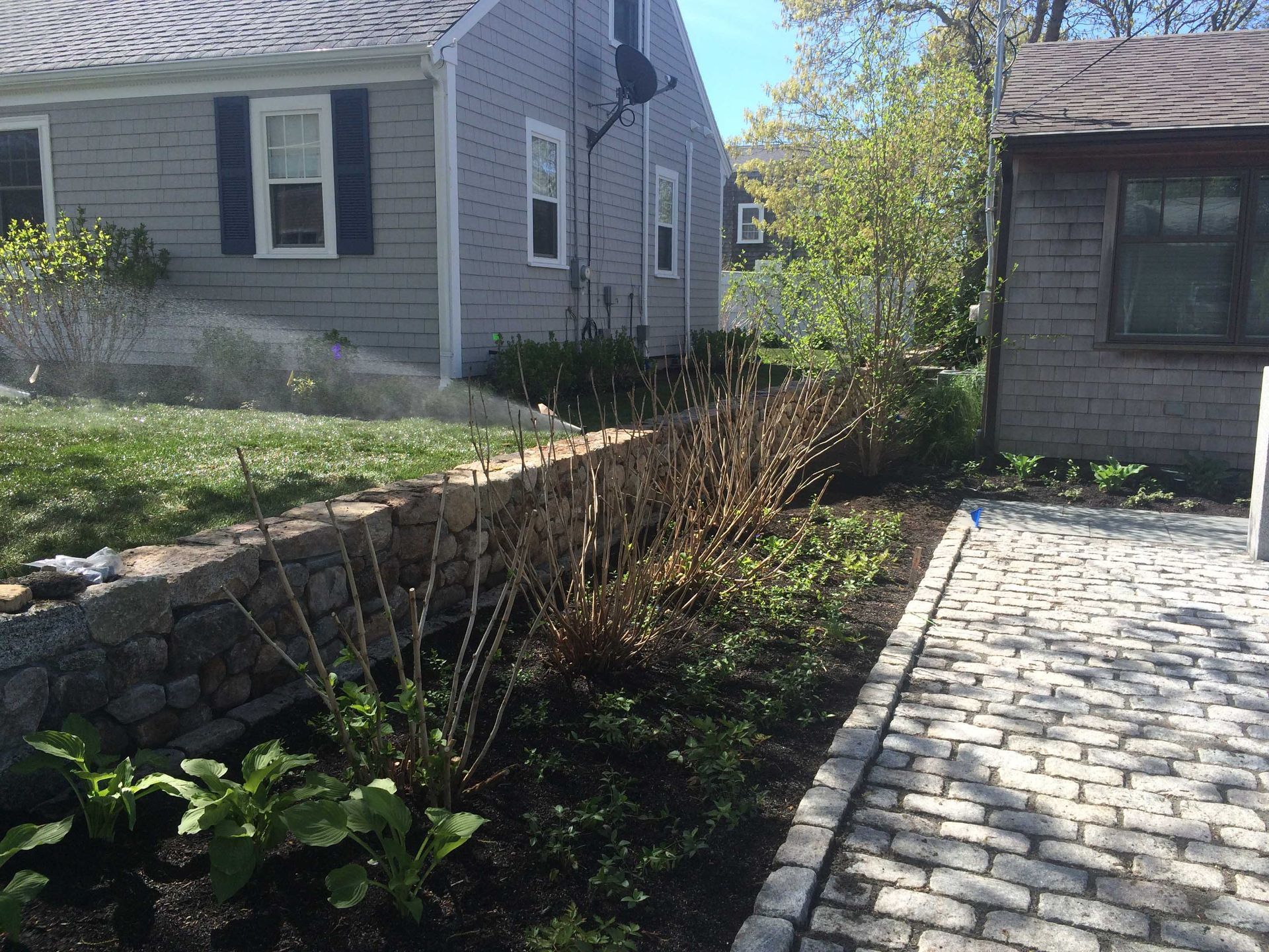 A gray house with a stone retaining wall and a cobblestone path lined with shrubs on a sunny day.