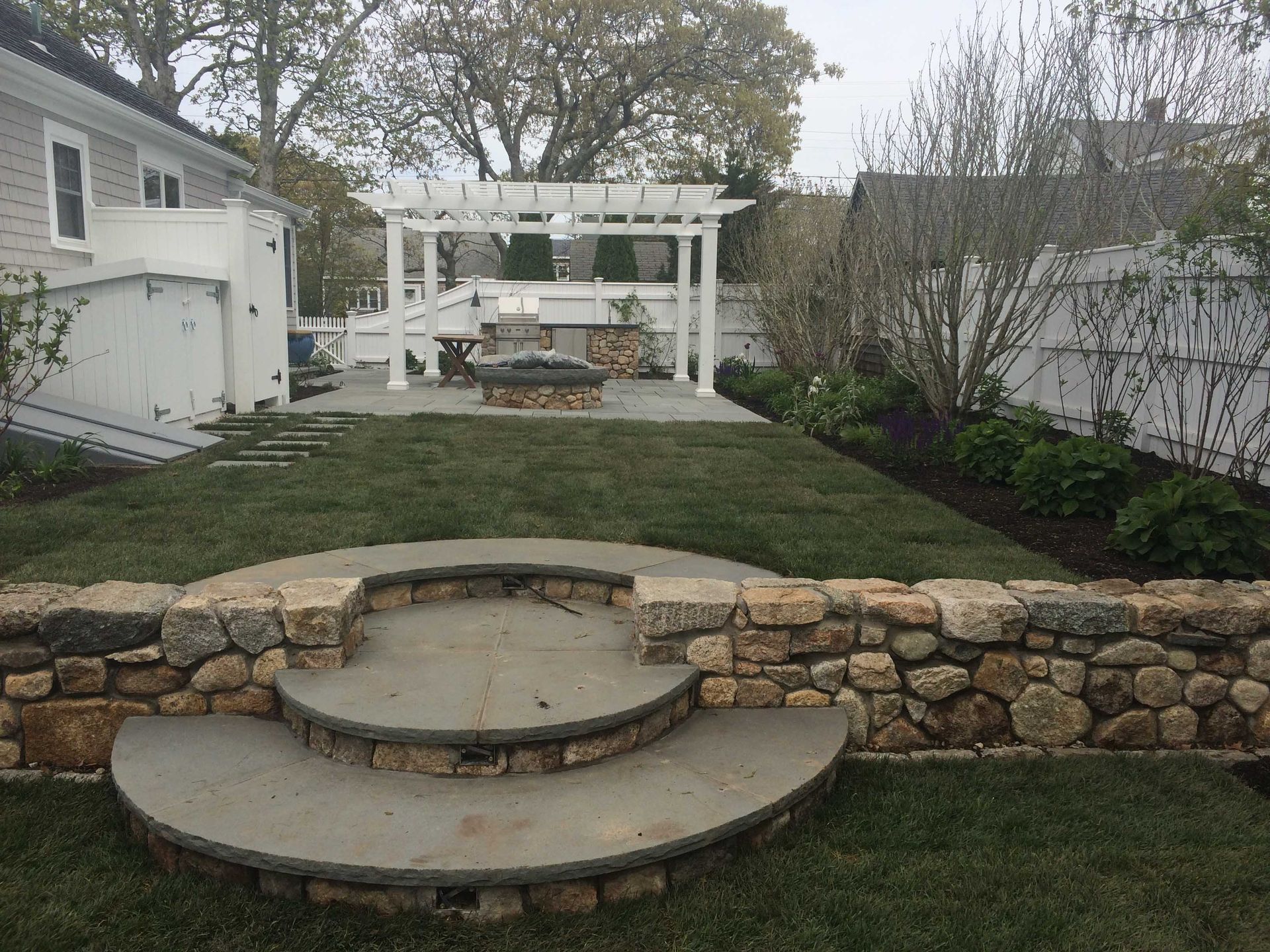A backyard with tiered stone steps, a lawn, a fire pit, and a white pergola under an overcast sky.