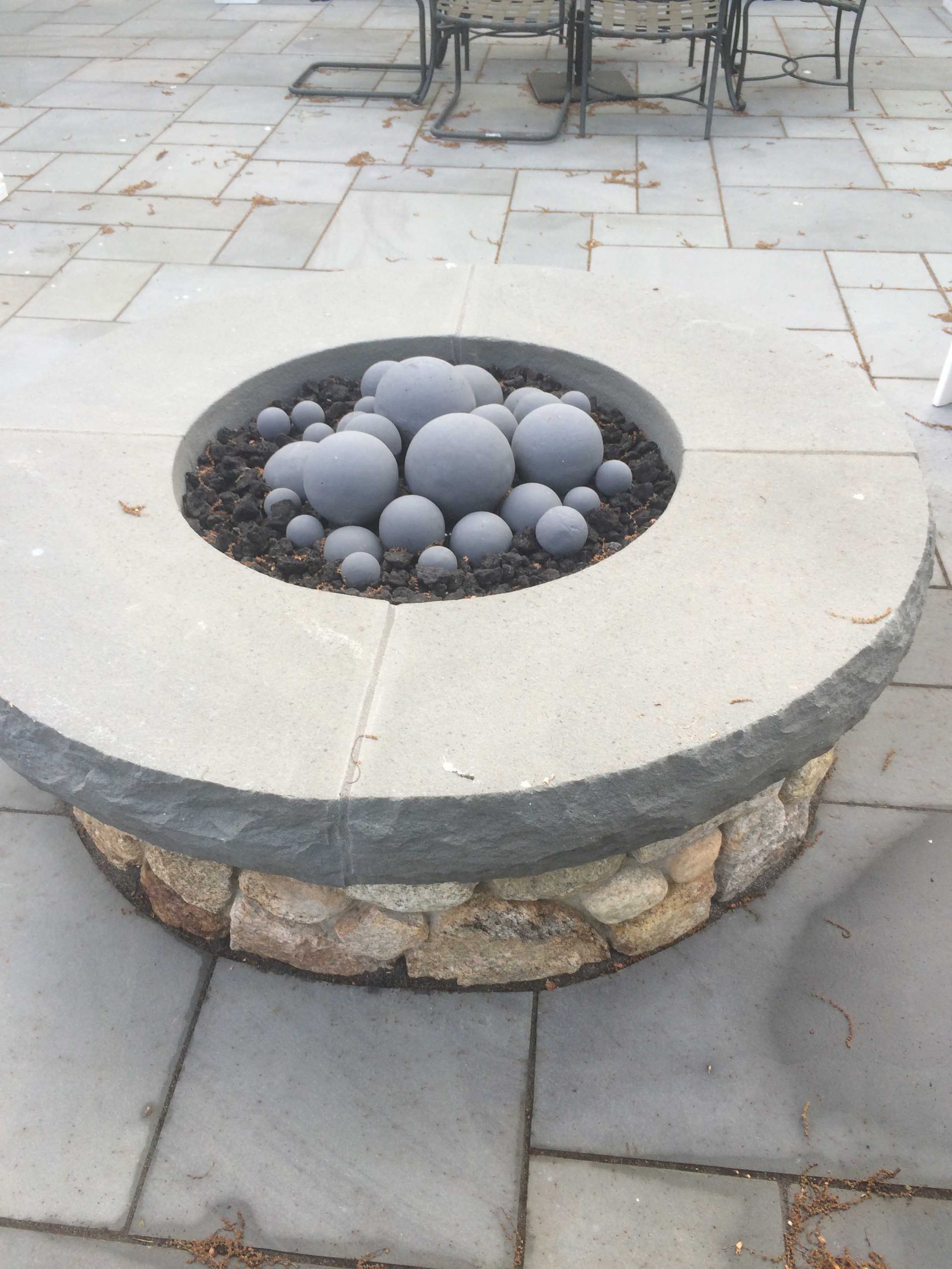 A circular stone fire pit sits on a patio, filled with black lava rock and various sizes of decorative gray concrete orbs.