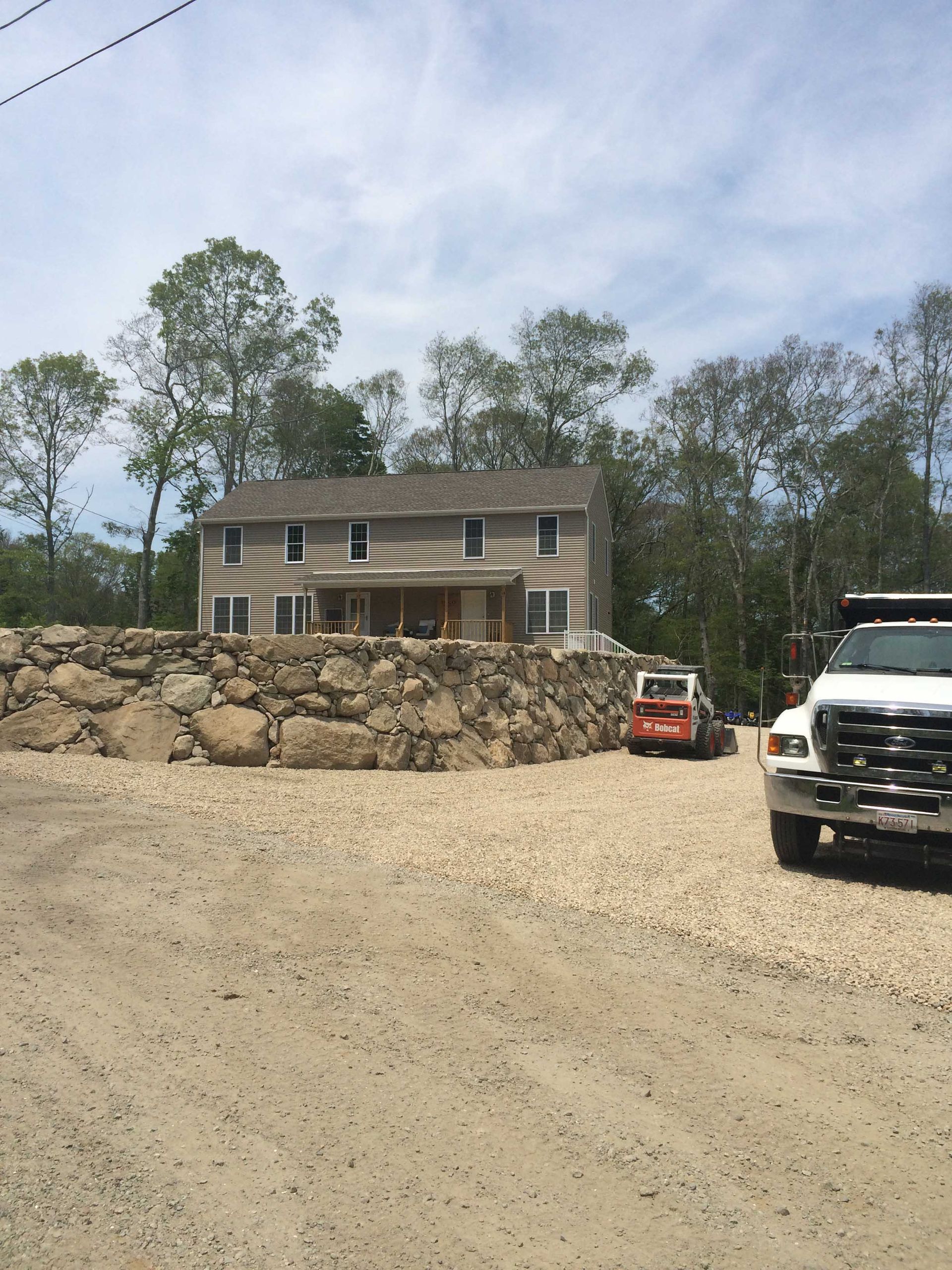 A two-story house sits atop a large stone retaining wall next to a parked dump truck and a small piece of construction gear.