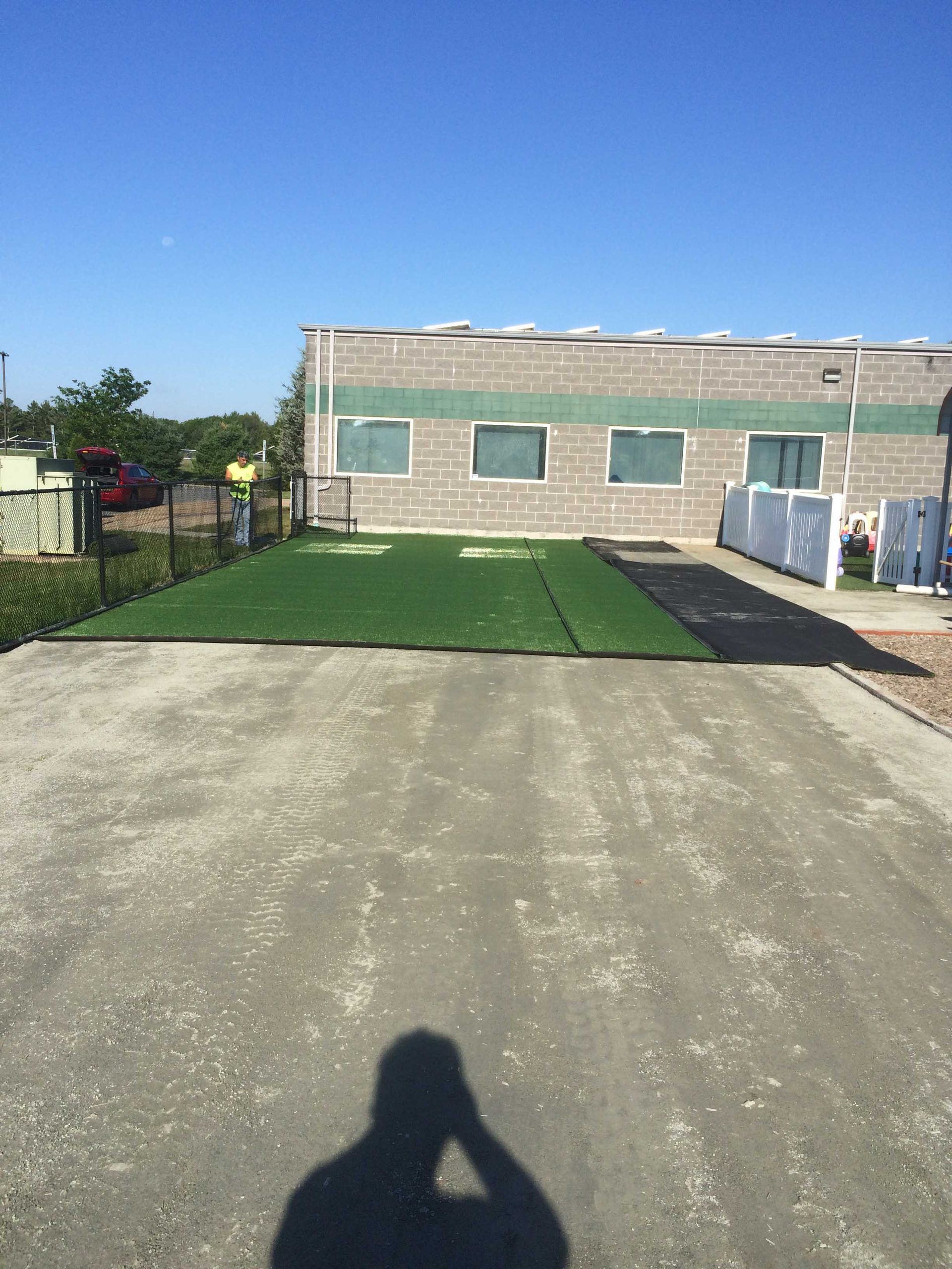 A strip of artificial turf sits on a gravel lot in front of a grey industrial building under a bright blue sky.