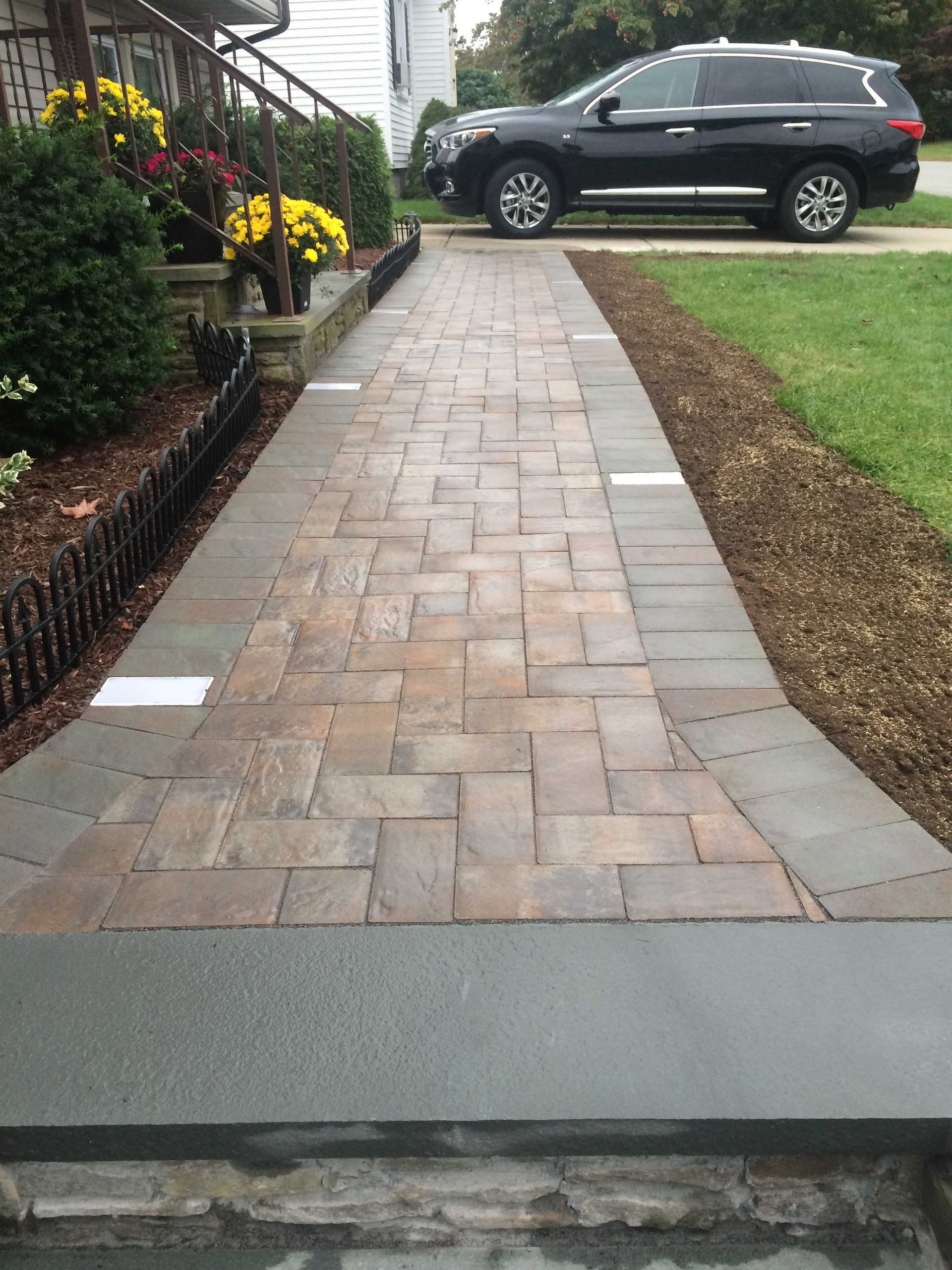 A paved walkway with a herringbone pattern in the center and dark border stones, leading toward a house and a parked SUV.