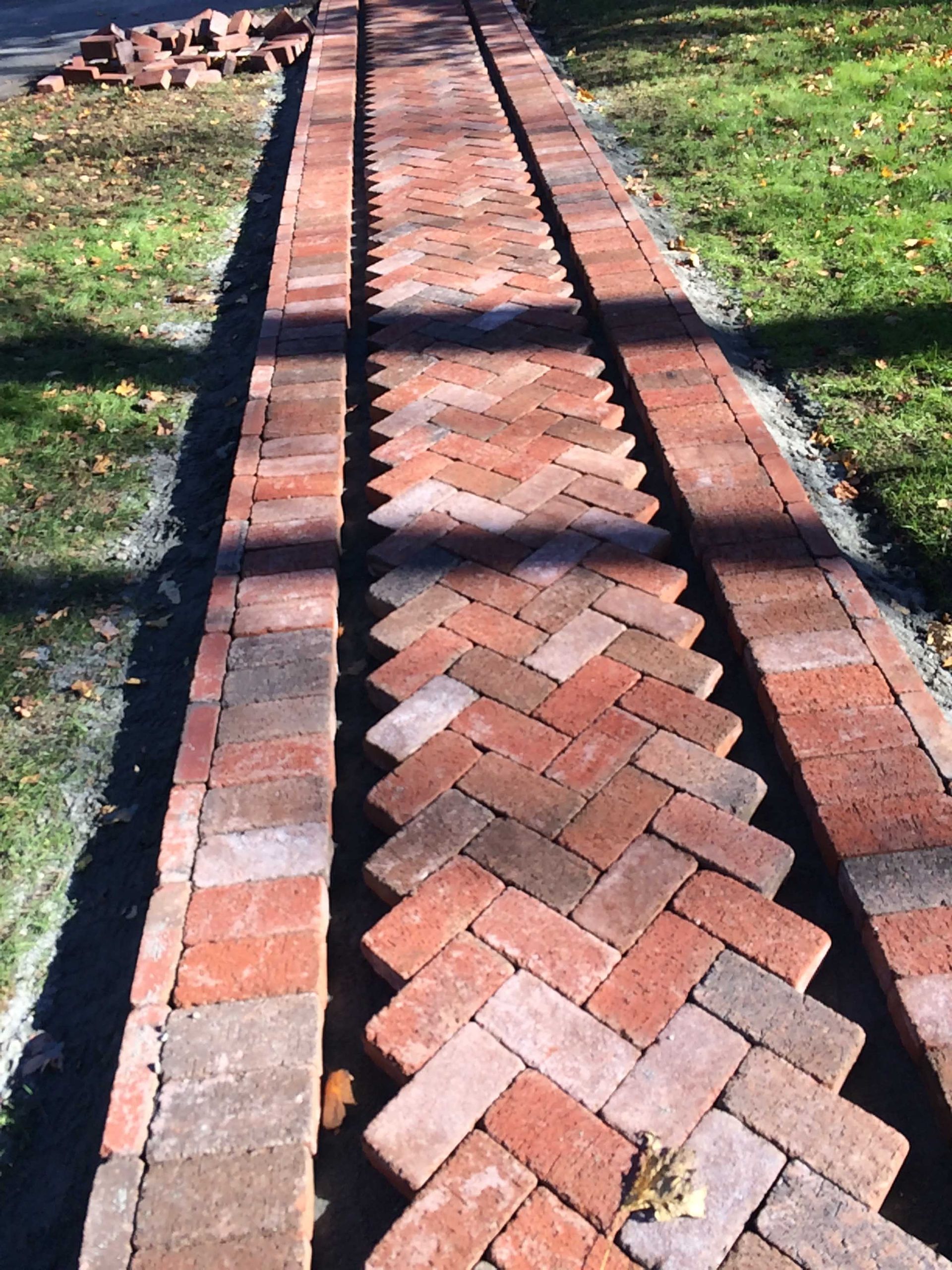 A partially constructed red brick walkway features a herringbone center pattern bordered by two straight rows of bricks.
