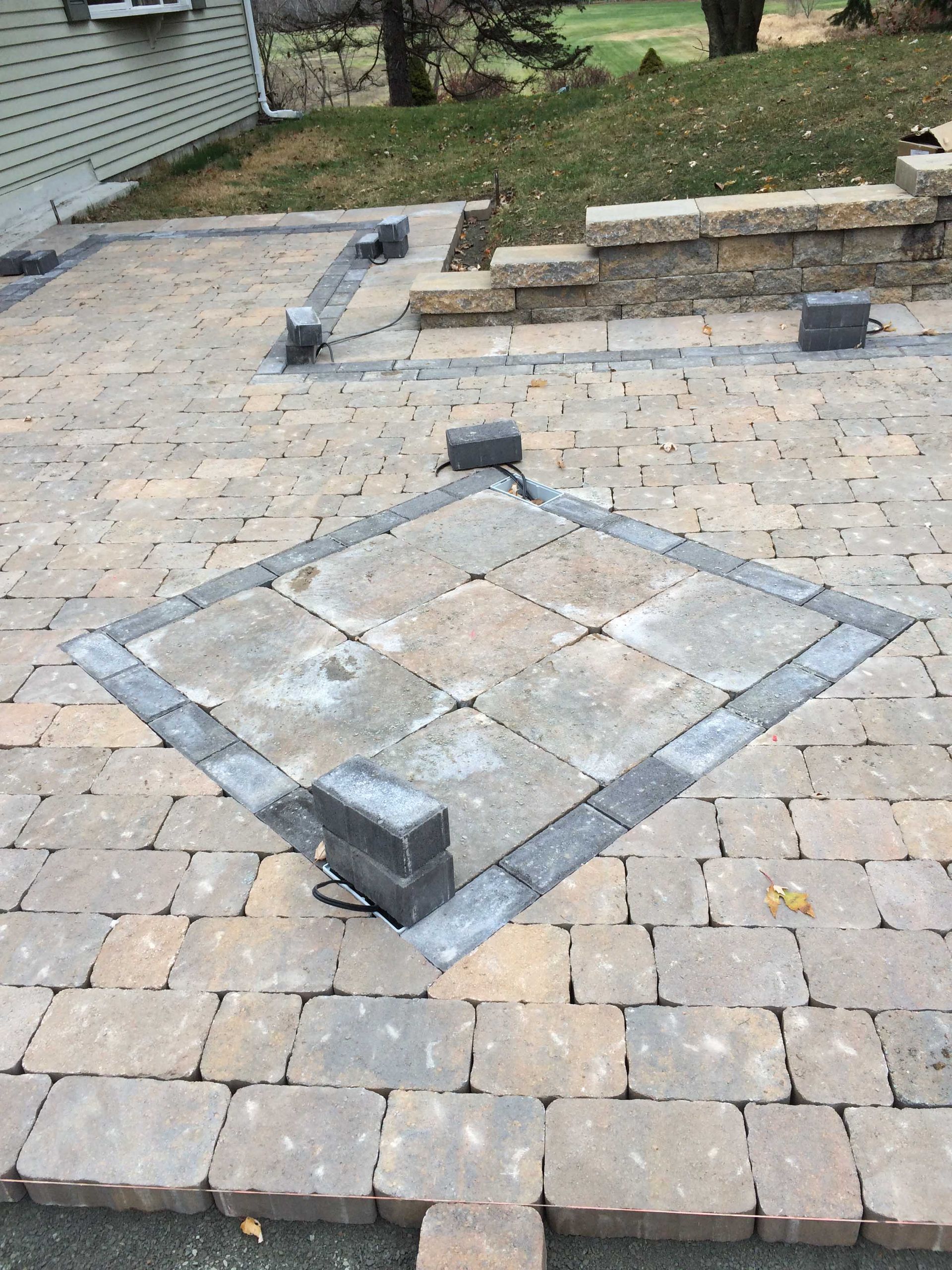 A square fire pit area under construction on a stone patio, with light-colored pavers framed by dark charcoal-grey bricks.