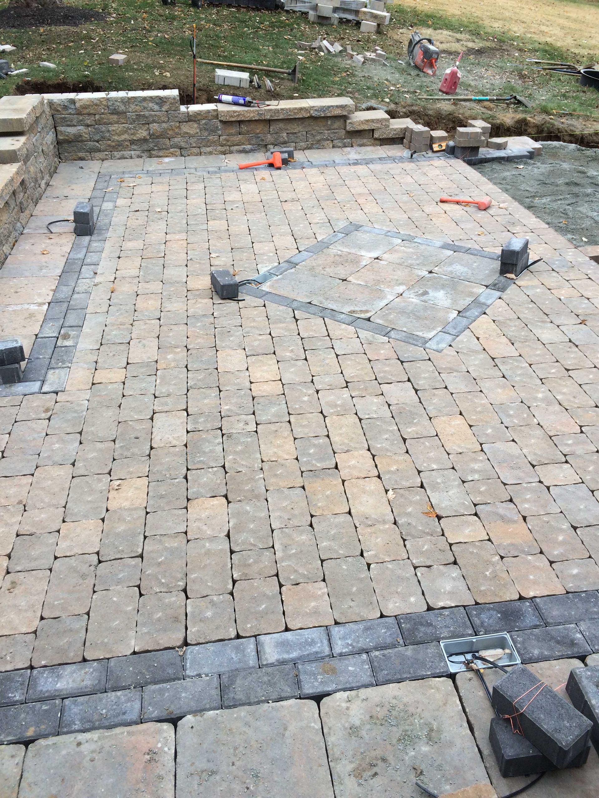 A newly laid outdoor stone patio with a patterned border and a square center design, partially surrounded by a stone wall.