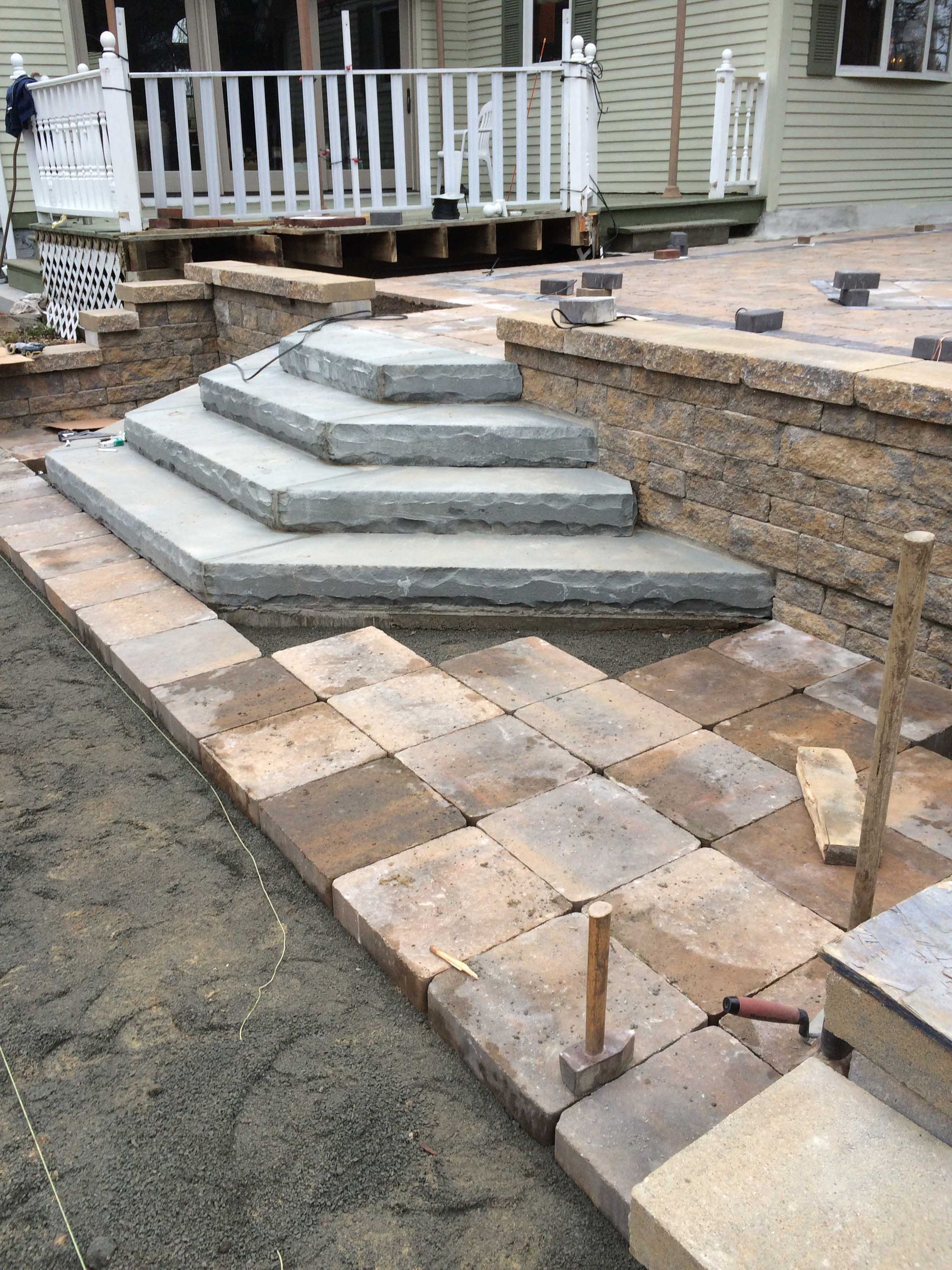 A stone staircase under construction leads to a house deck, with partially laid pavers in the foreground on a dirt base.