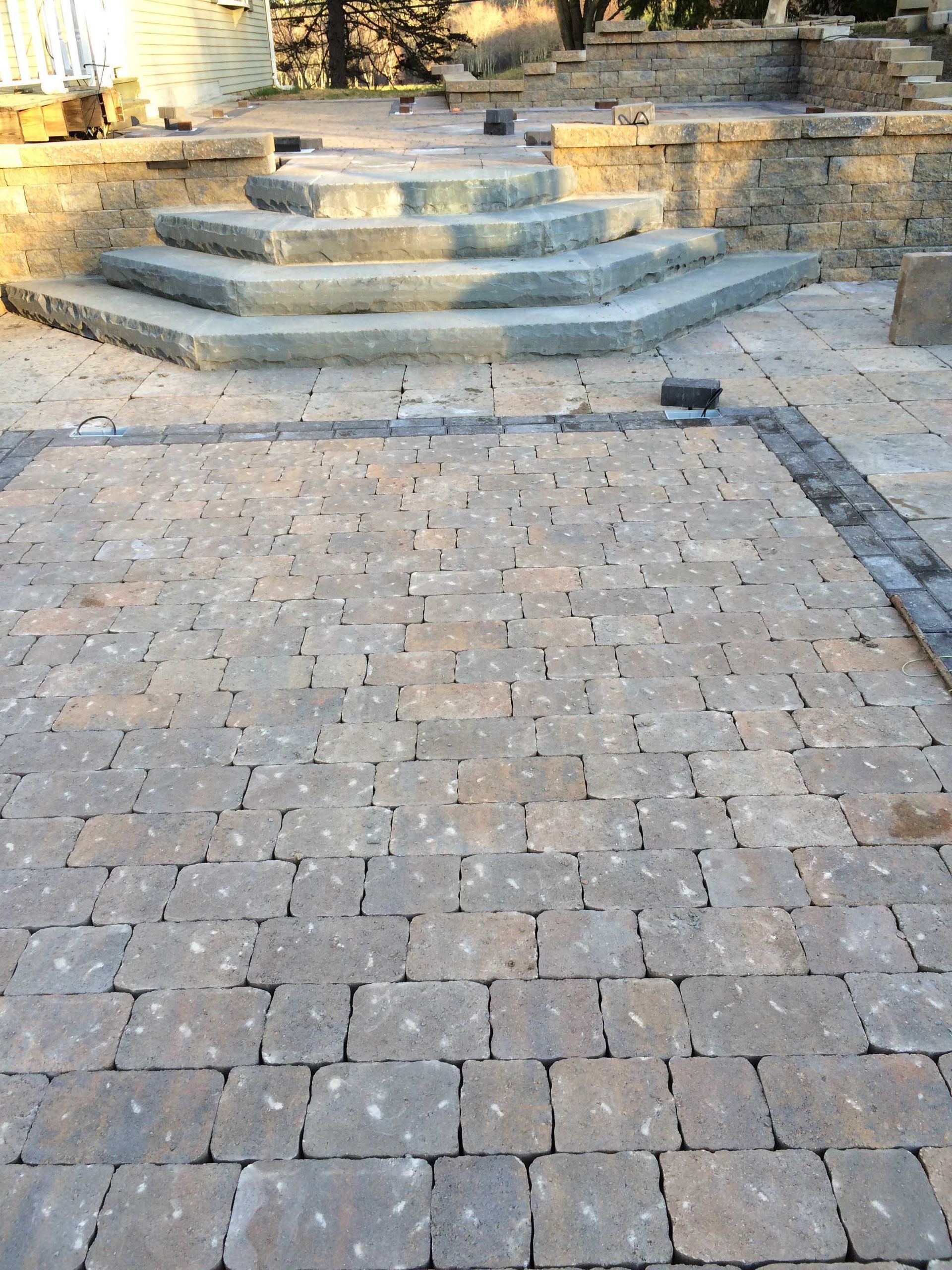 A stone patio with a textured gray paver pattern leading up to three curved stone steps in a residential backyard.