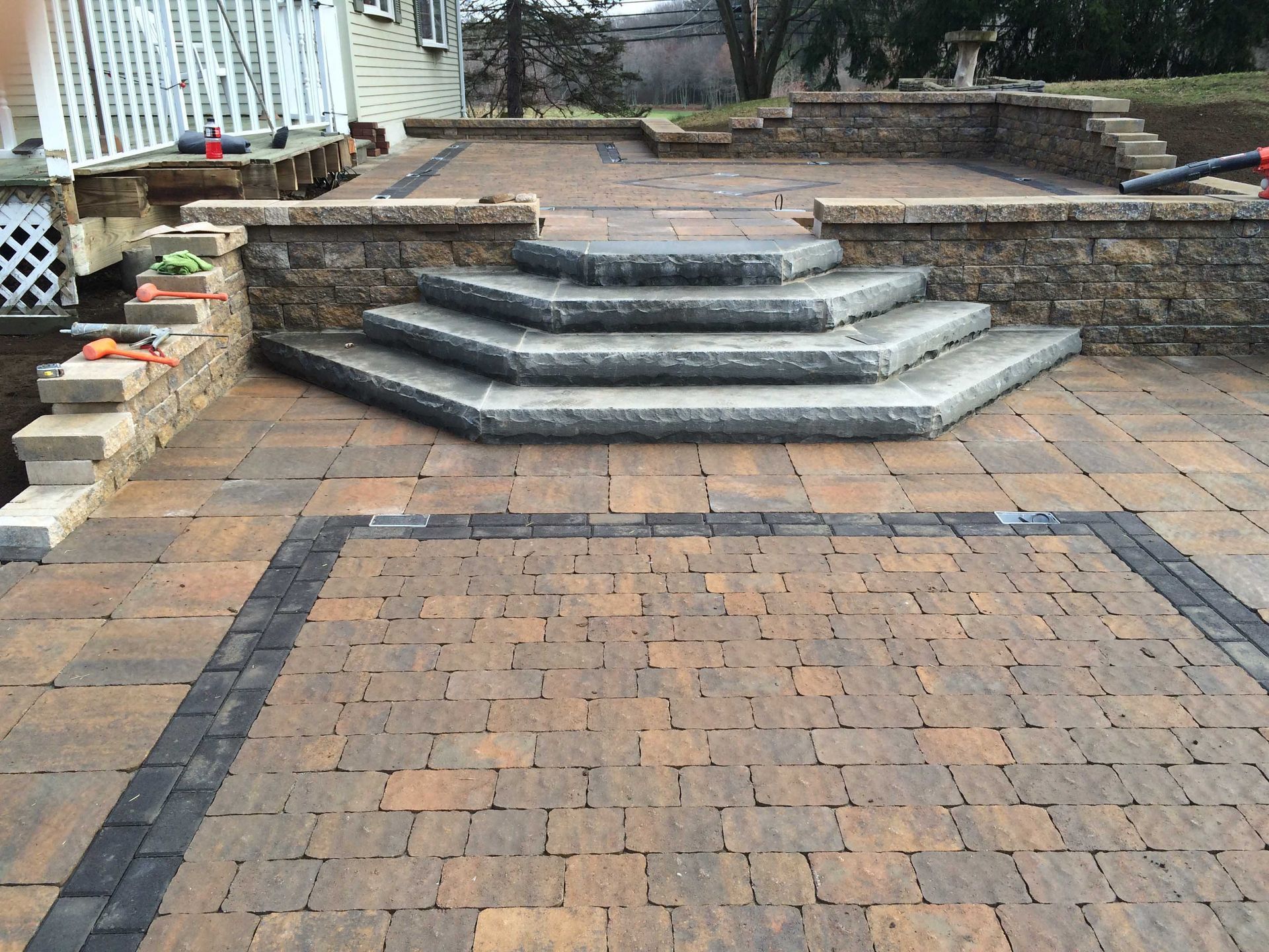 A tiered paver patio with a stone retaining wall and three wide, gray, hexagonal-shaped steps leading to an upper level.