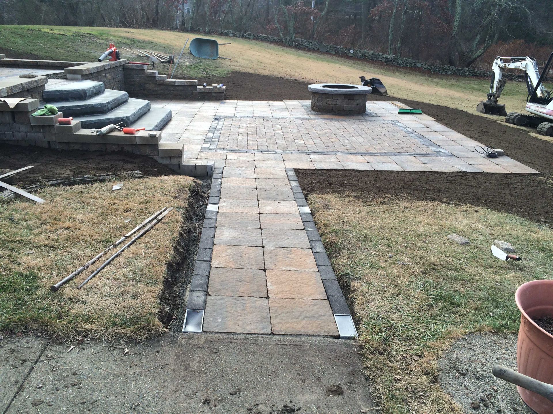 A new paver walkway and patio with a fire pit under construction in a backyard, with steps to the left and heavy equipment.