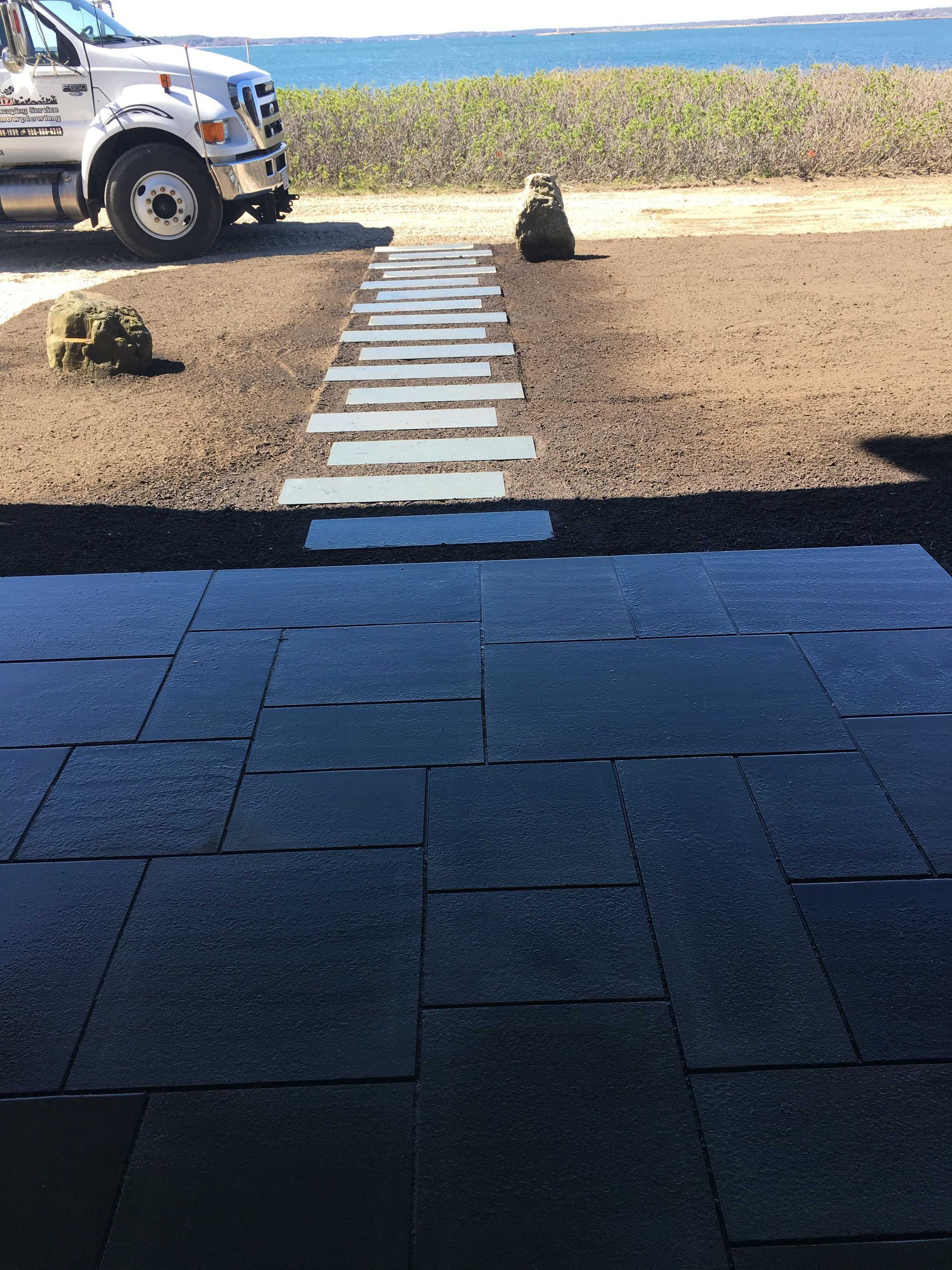 A stone walkway leads from a dark patio toward a white truck and a grassy area near the water.
