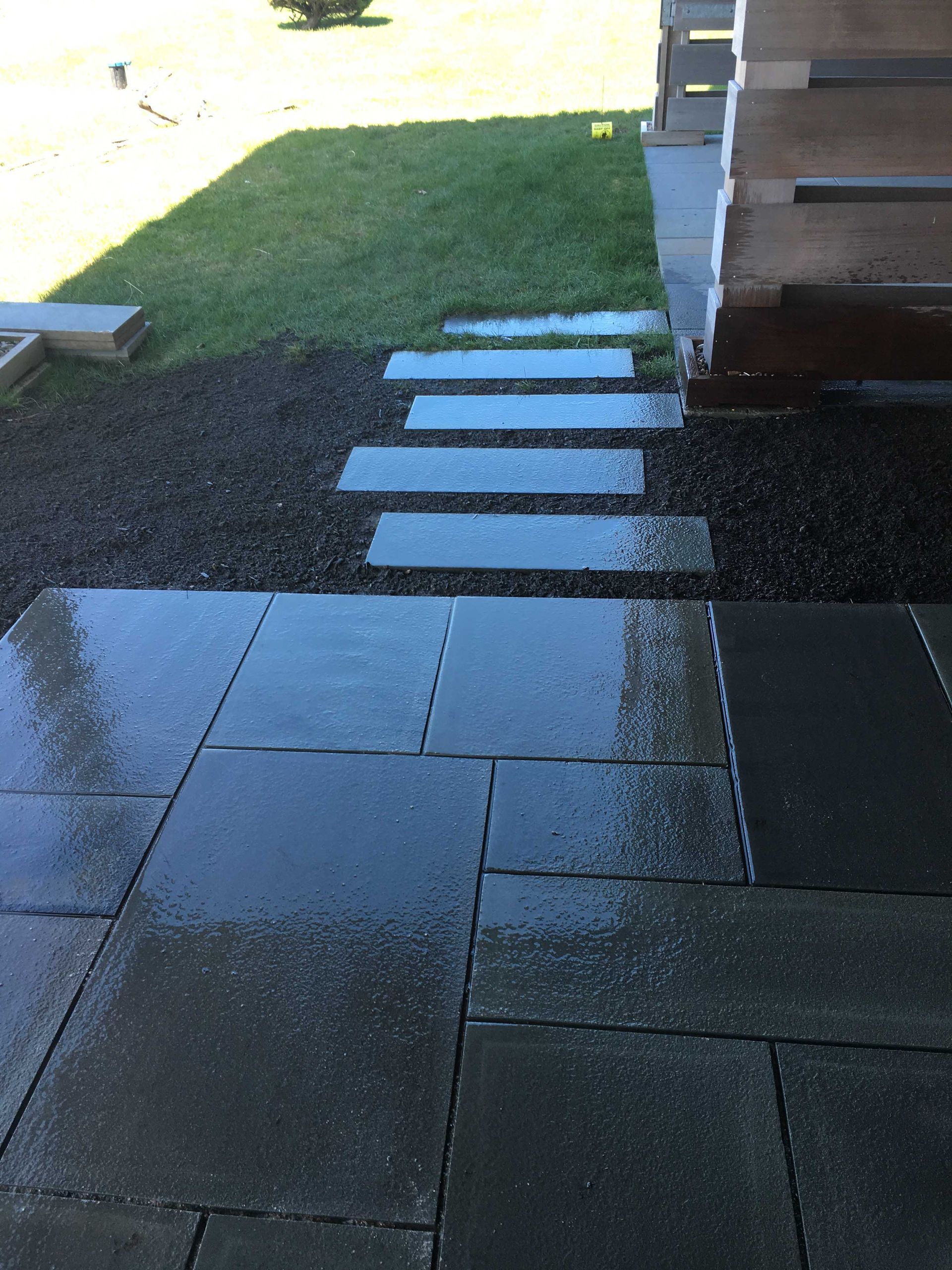Wet, dark stone patio leading to a set of stone steps surrounded by gravel and grass near a building.