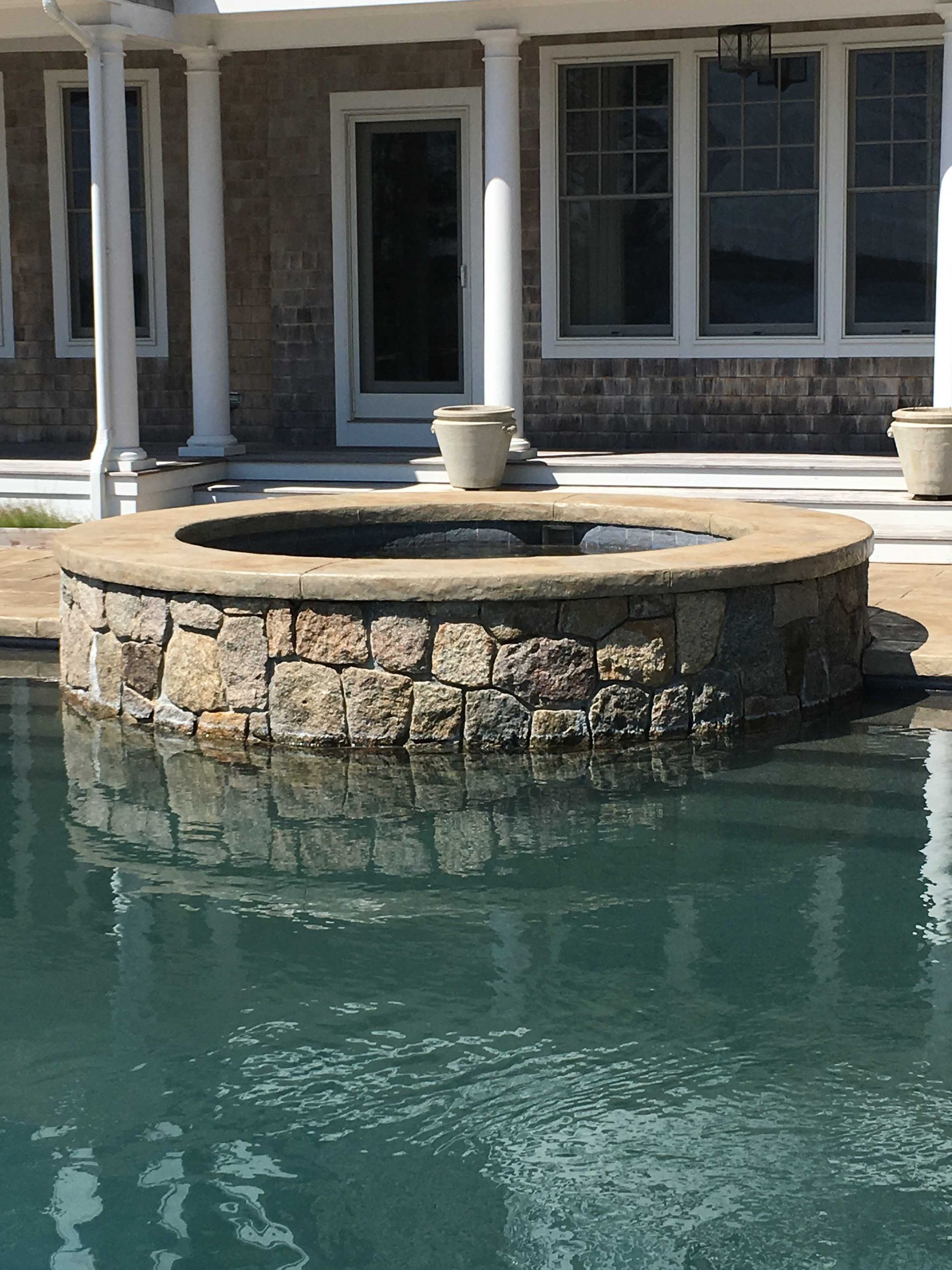 A round stone hot tub sits partially submerged in a pool in front of a house with white columns and large windows.