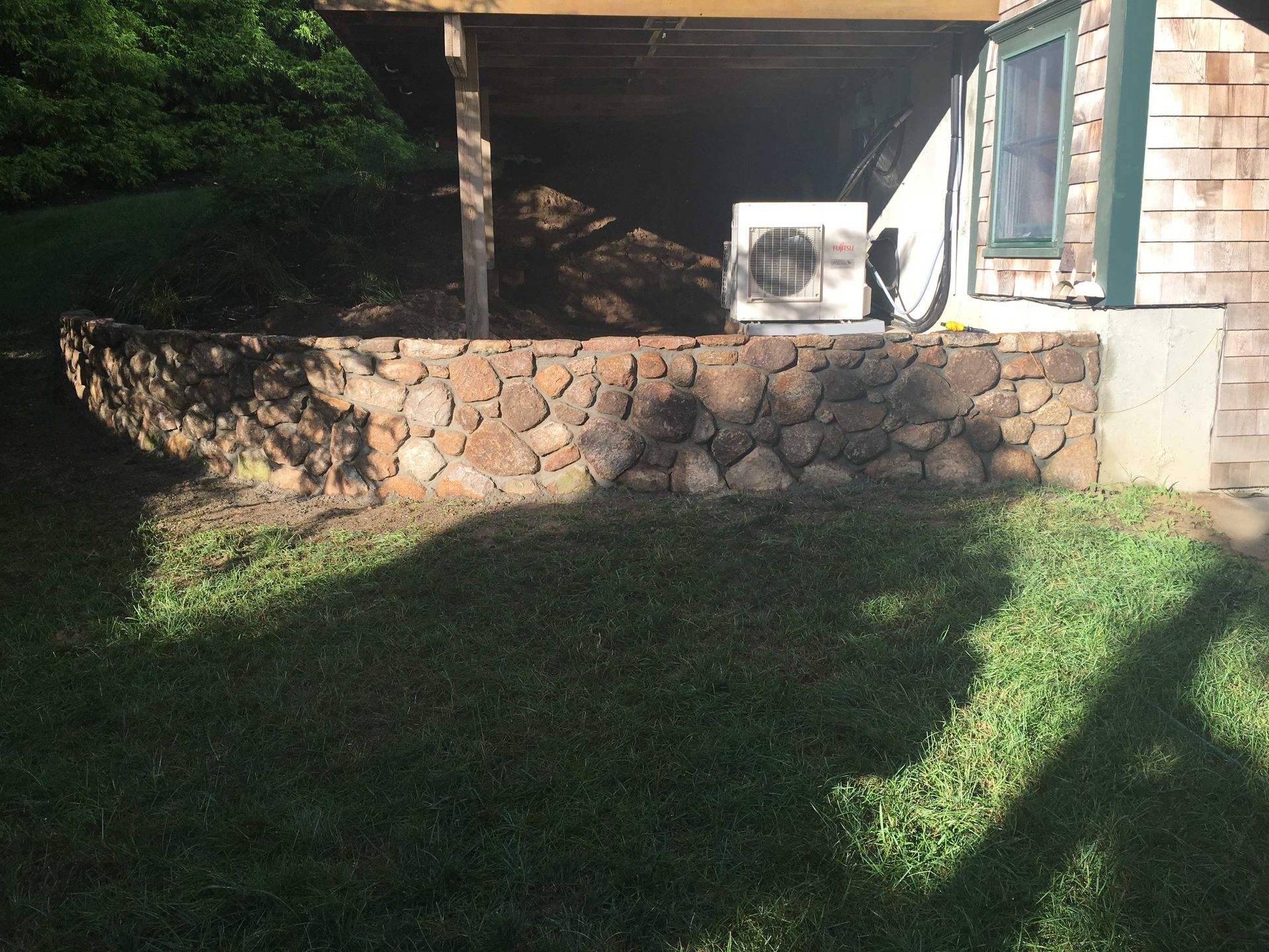 A stone retaining wall built in front of a shaded porch area with a white HVAC unit near a house with wood siding.
