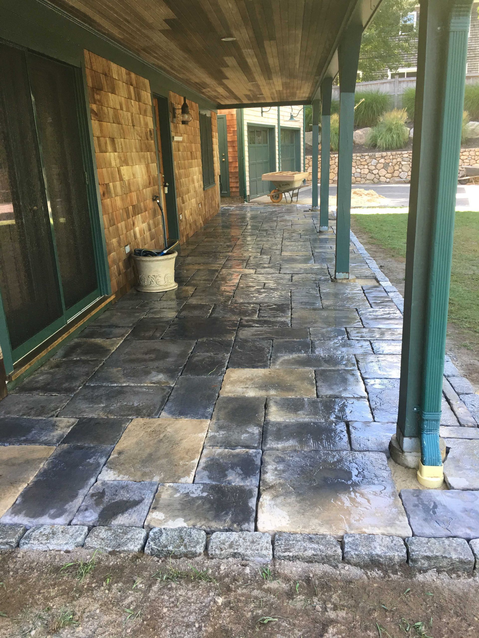 A stone patio with dark, rectangular pavers under a covered porch attached to a tan brick wall with a sliding glass door.