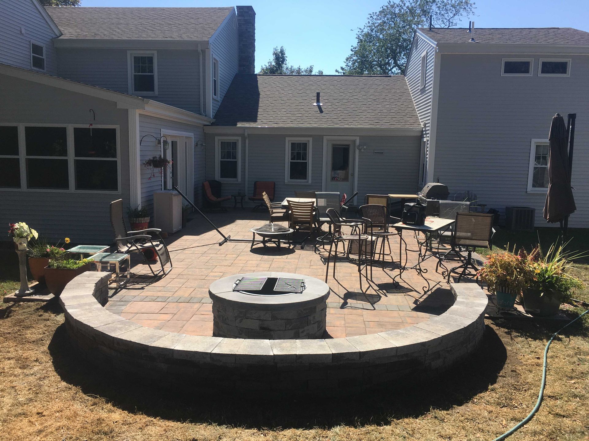 A backyard patio featuring a stone fire pit, a curved stone seating wall, dining furniture, and light gray siding.