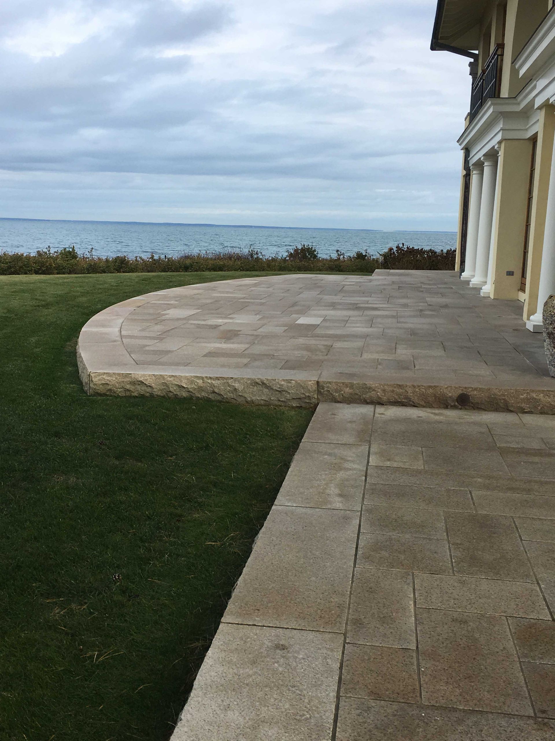 A stone patio overlooking a body of water next to a house with white columns under a cloudy sky.