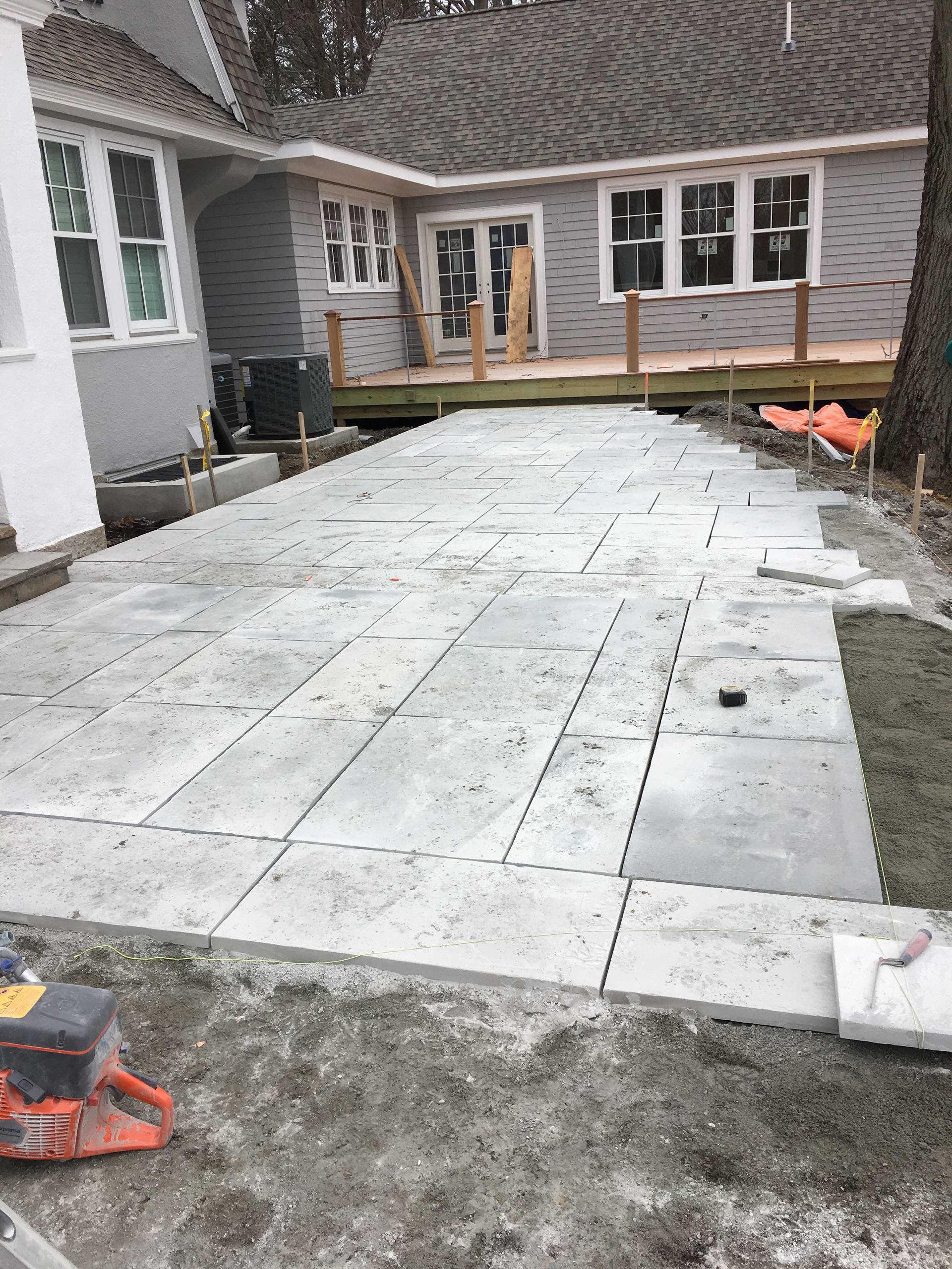 Gray stone patio being installed in a backyard next to a house with a wooden deck.