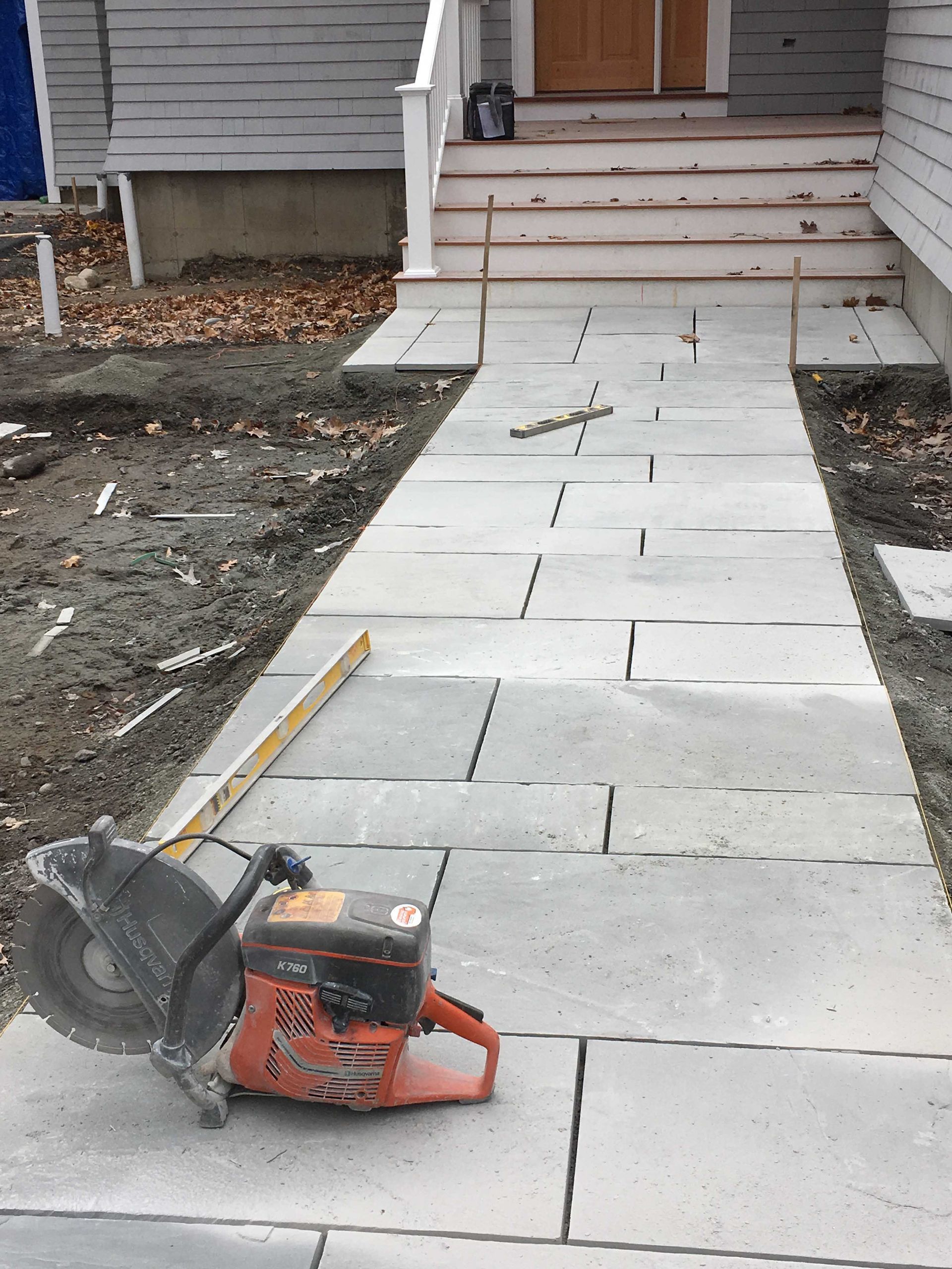 A gas-powered masonry saw sits on a newly laid gray stone walkway leading to a house entrance.