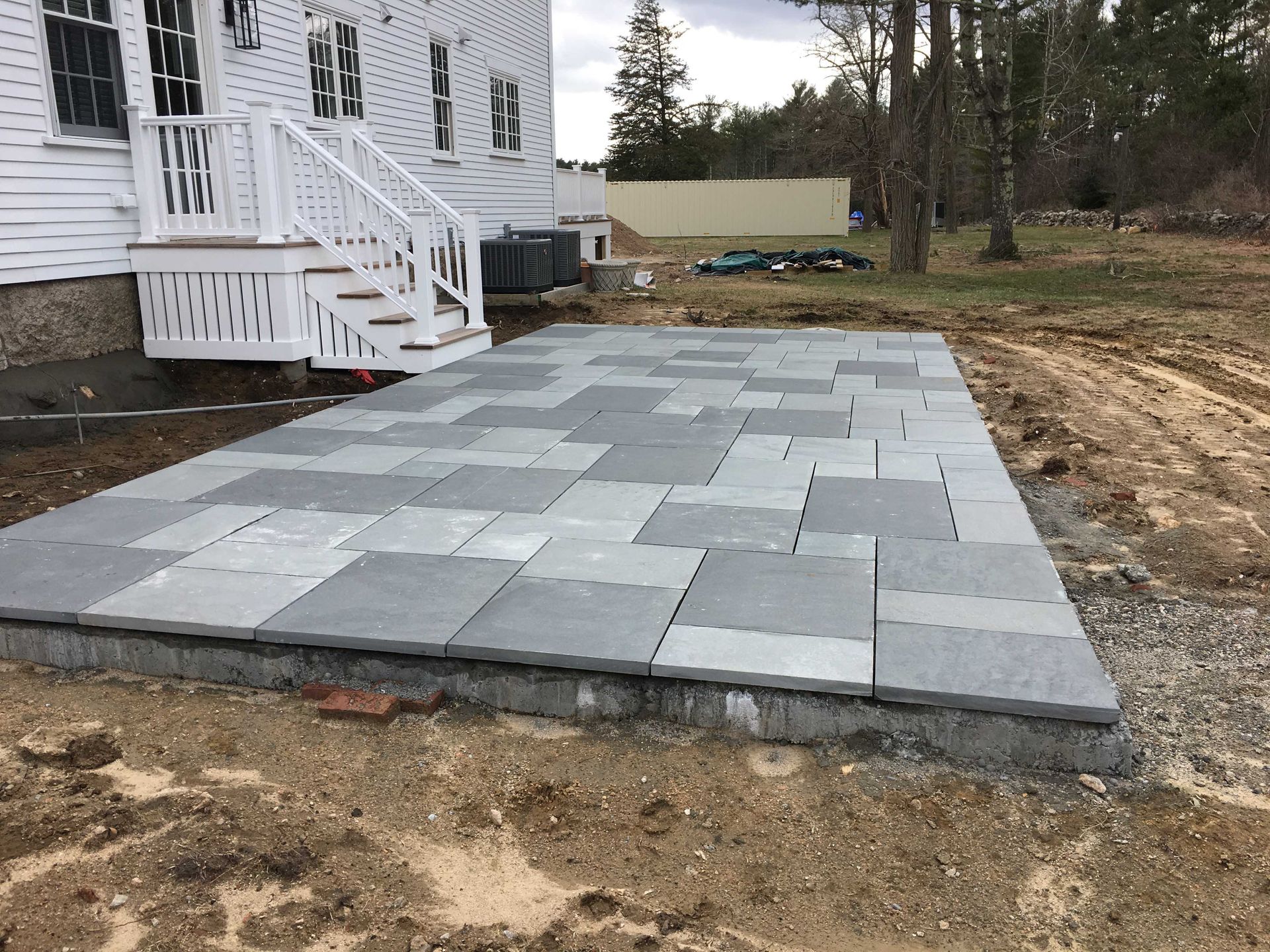 A new gray stone patio sits adjacent to a white house with a set of white stairs leading to the back door.