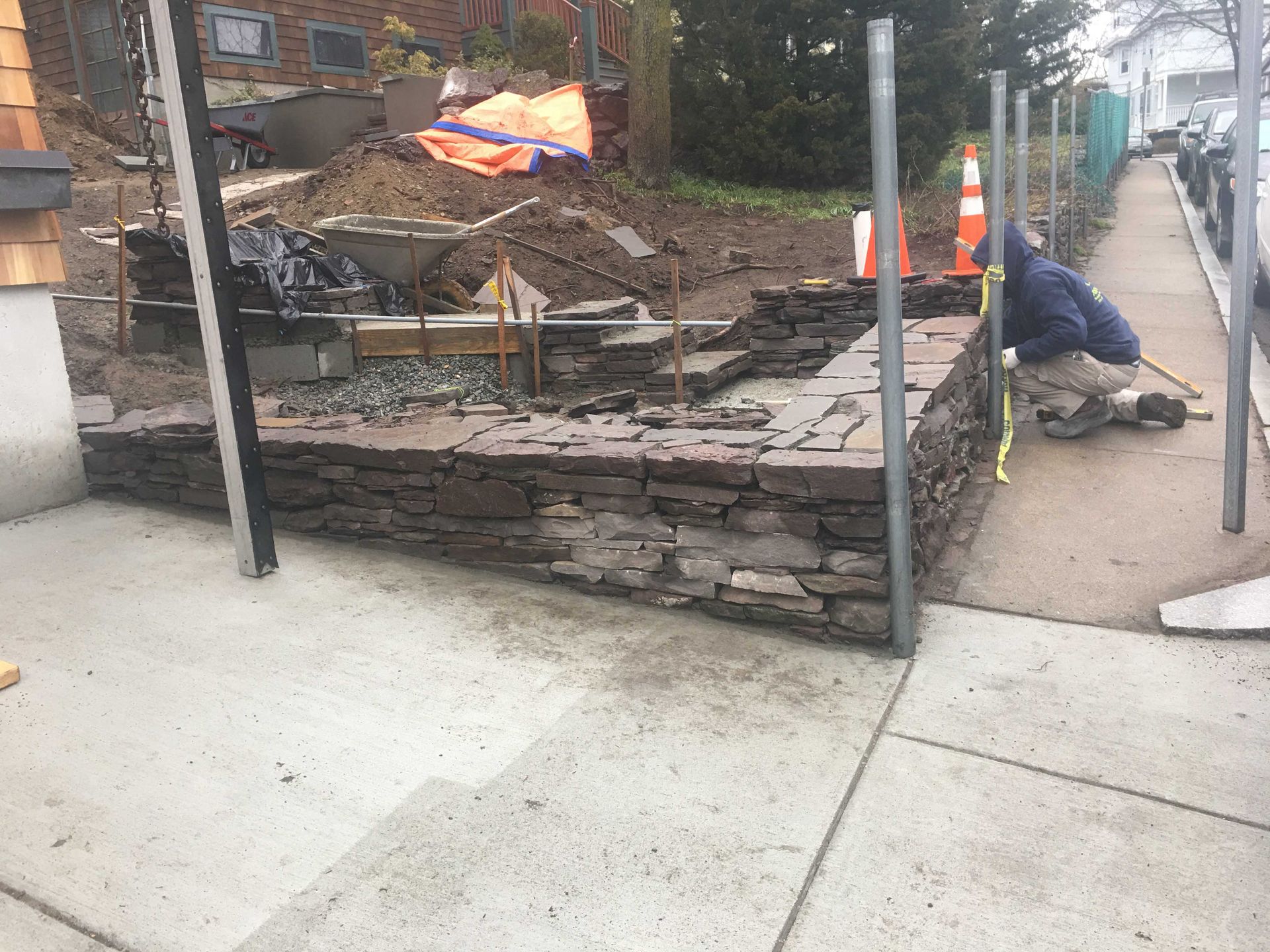 A construction worker installs metal fence posts next to a newly built stone retaining wall along a sidewalk.