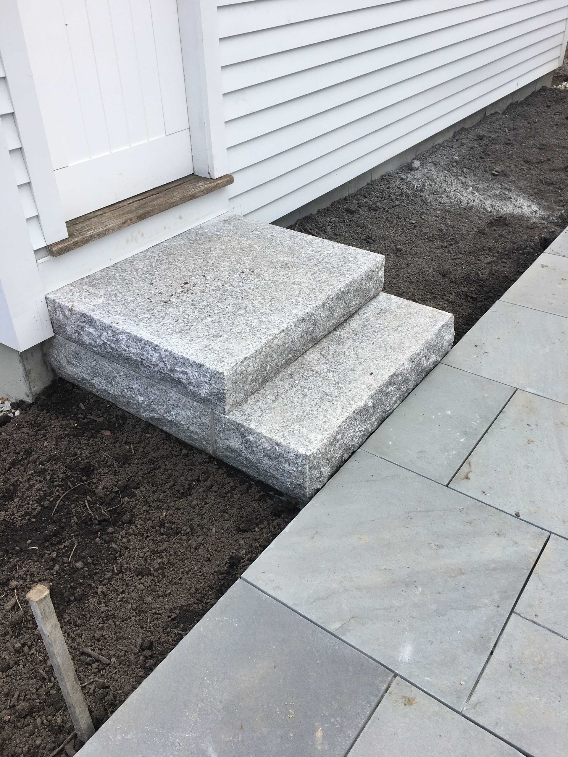 Two granite steps leading to a white door next to a patio of gray stone pavers and an area of dark soil.
