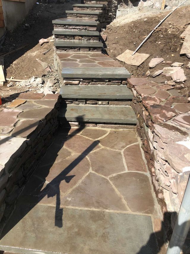 A stone pathway with a set of stairs leading upward through a construction site with exposed dirt and stacked stone walls.