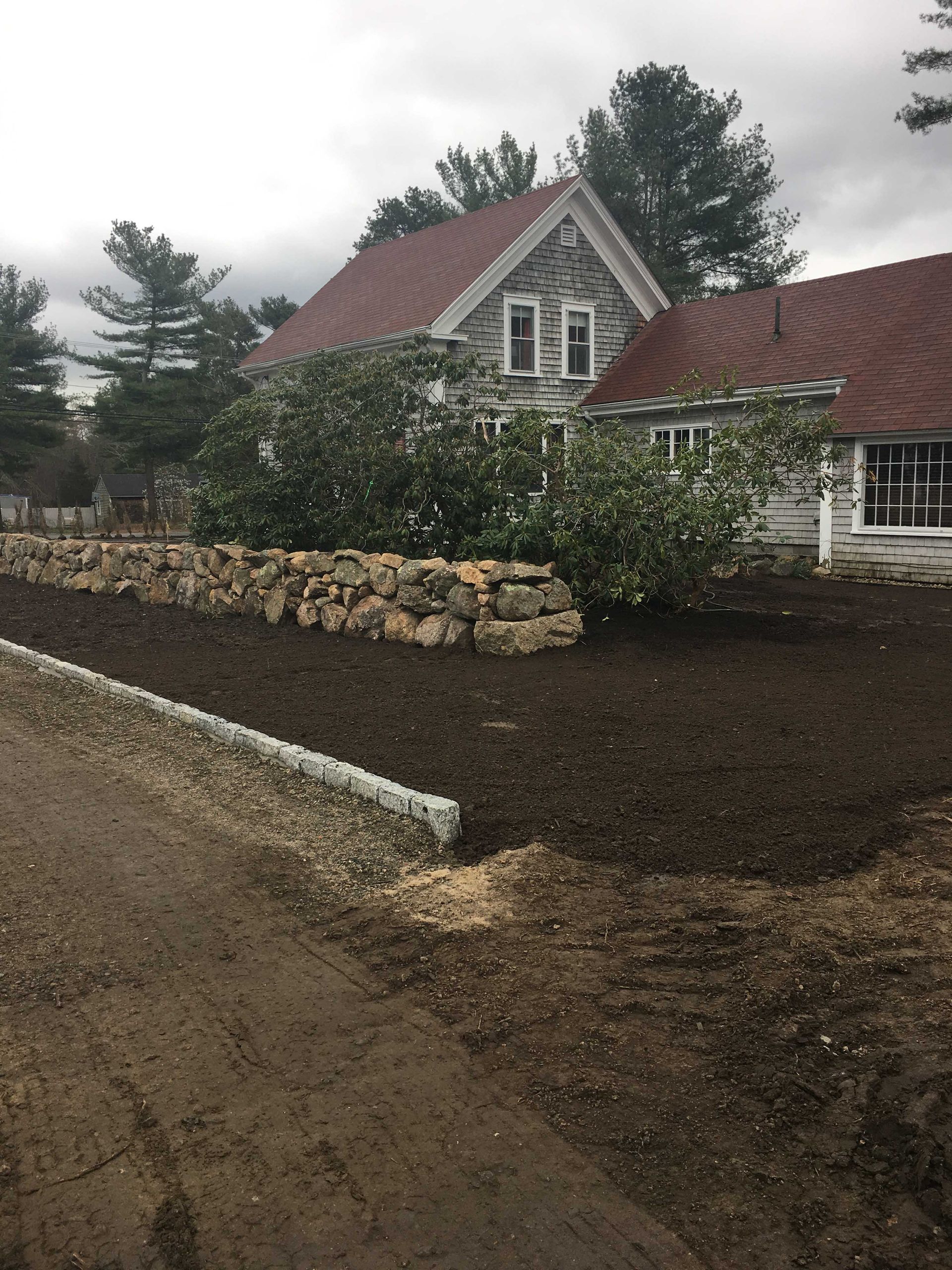 A stone house with a red roof stands behind a fresh mulch garden bed bordered by a low stone wall and a gravel path.