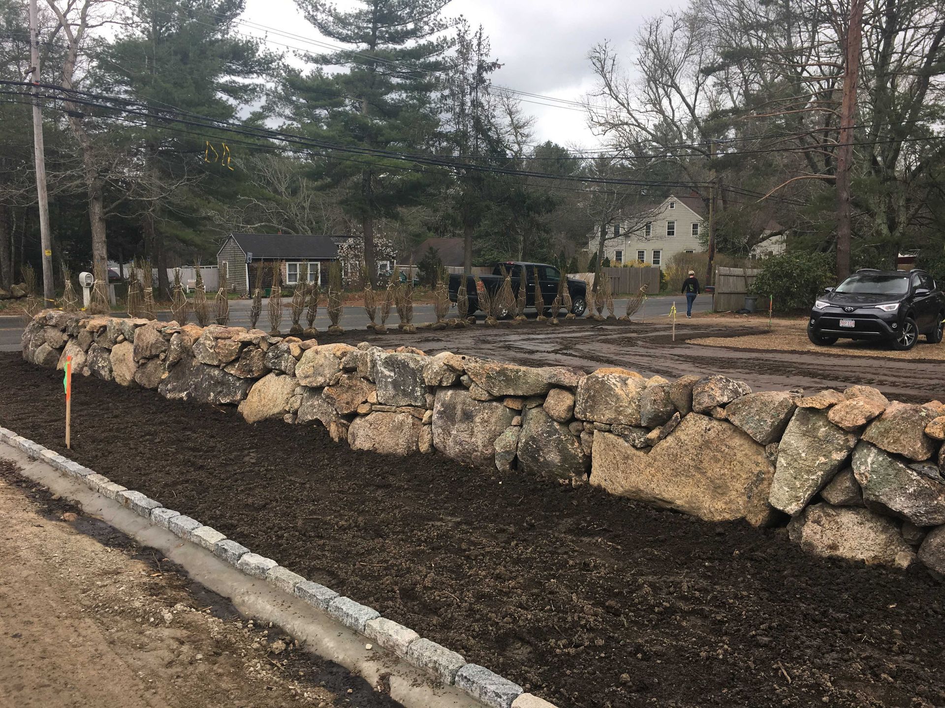 A newly constructed fieldstone wall sits behind a paved curb and fresh mulch, with trees and a house in the background.