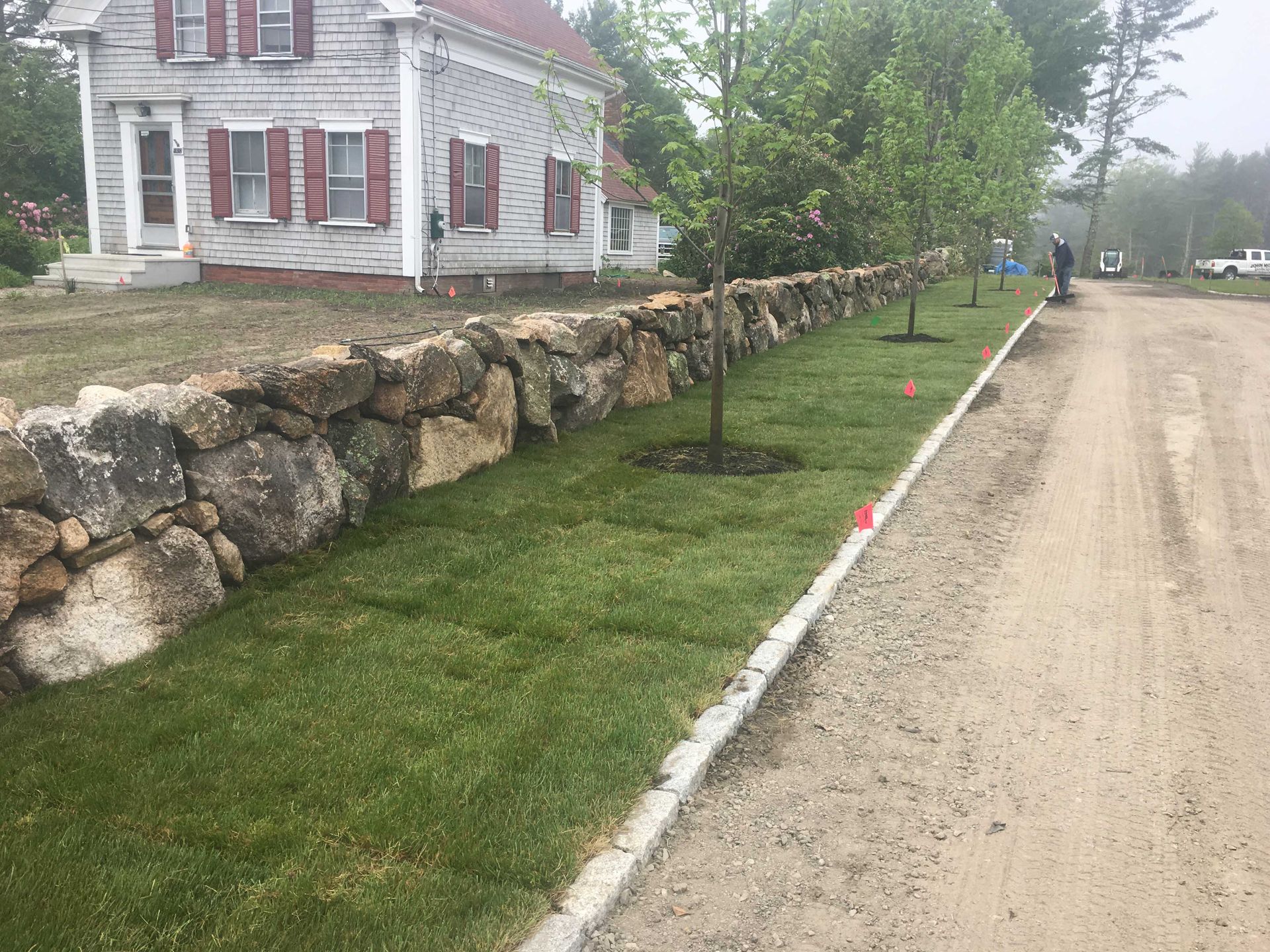 A stone wall runs along a dirt road, next to a house with white siding, red shutters, and newly planted young trees.