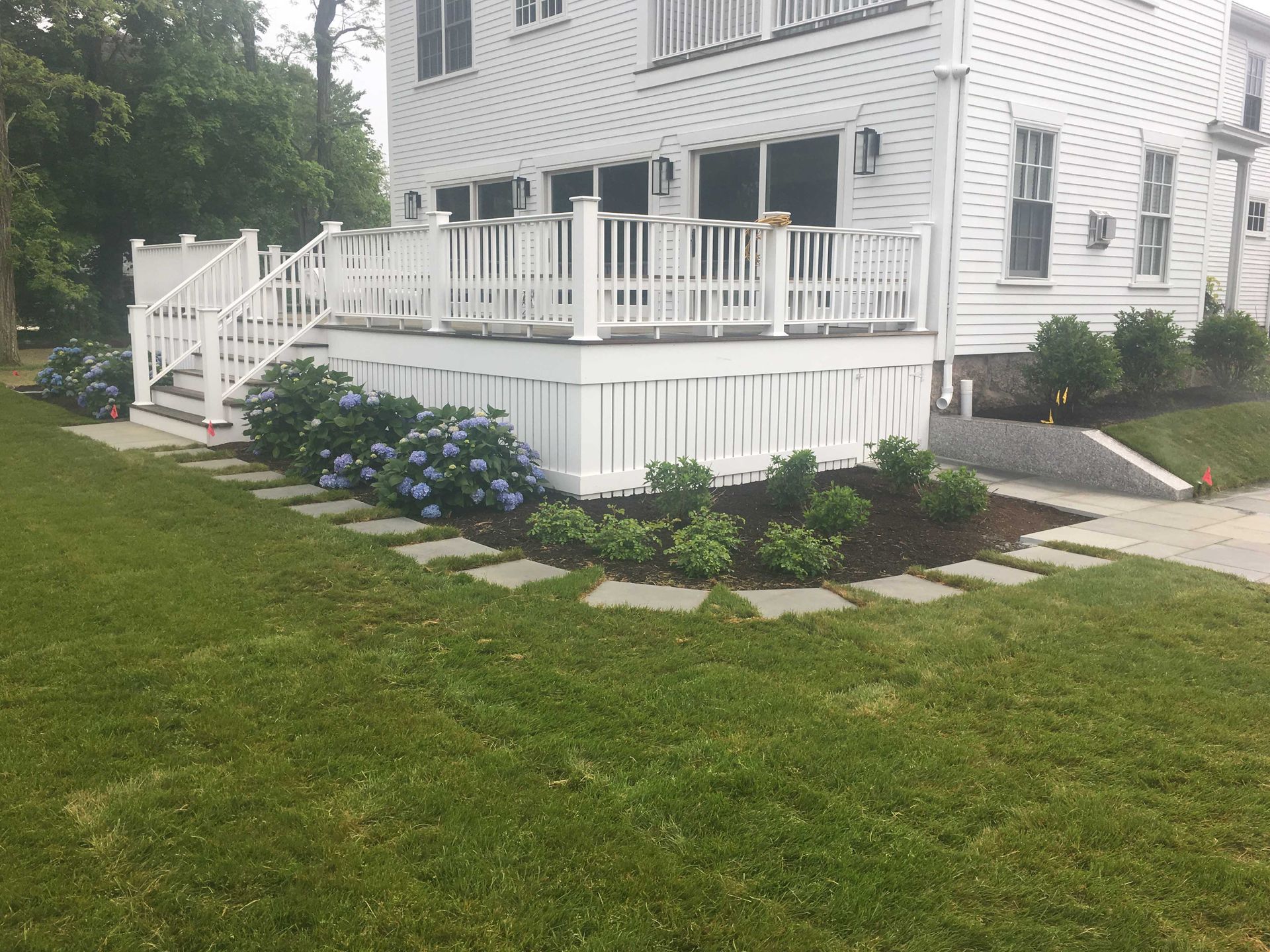 White house with a raised wooden deck, surrounded by hydrangea bushes and stone pavers in a grassy yard.