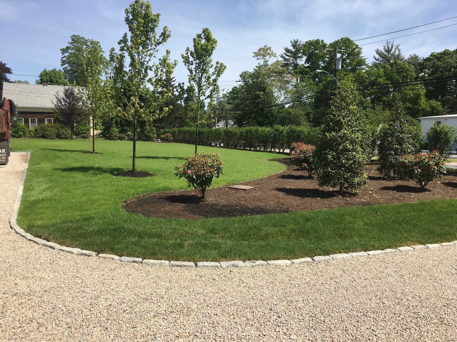 A neatly landscaped front lawn with trees and shrubs bordering a curved gravel driveway, under a clear blue sky.