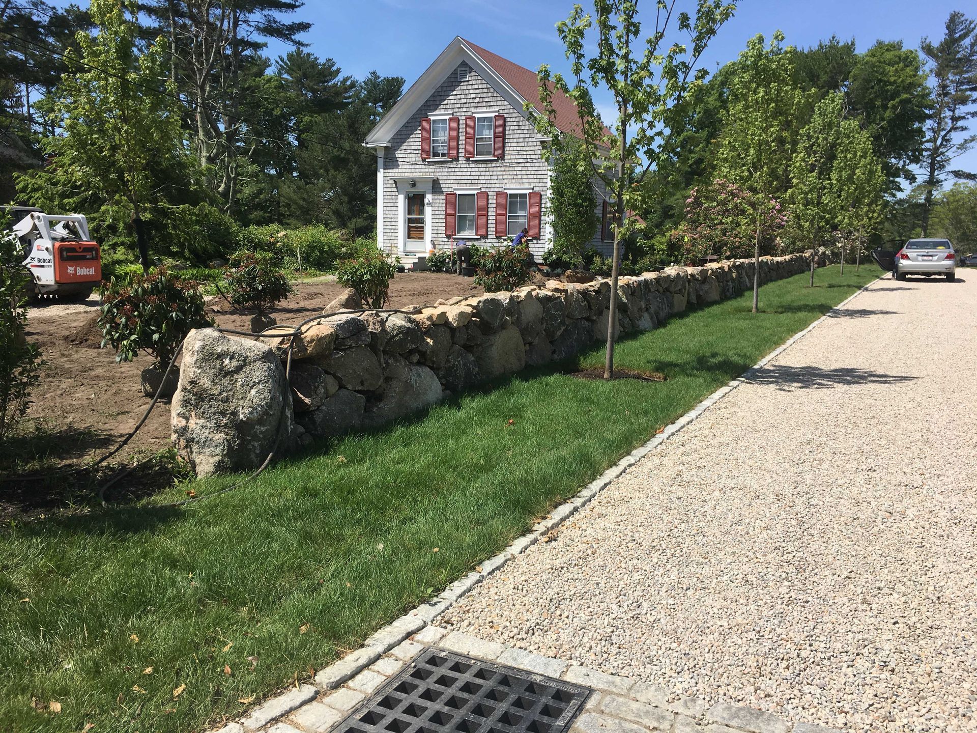 A stone wall borders a gravel driveway in front of a white house with red shutters and a green lawn under a sunny sky.