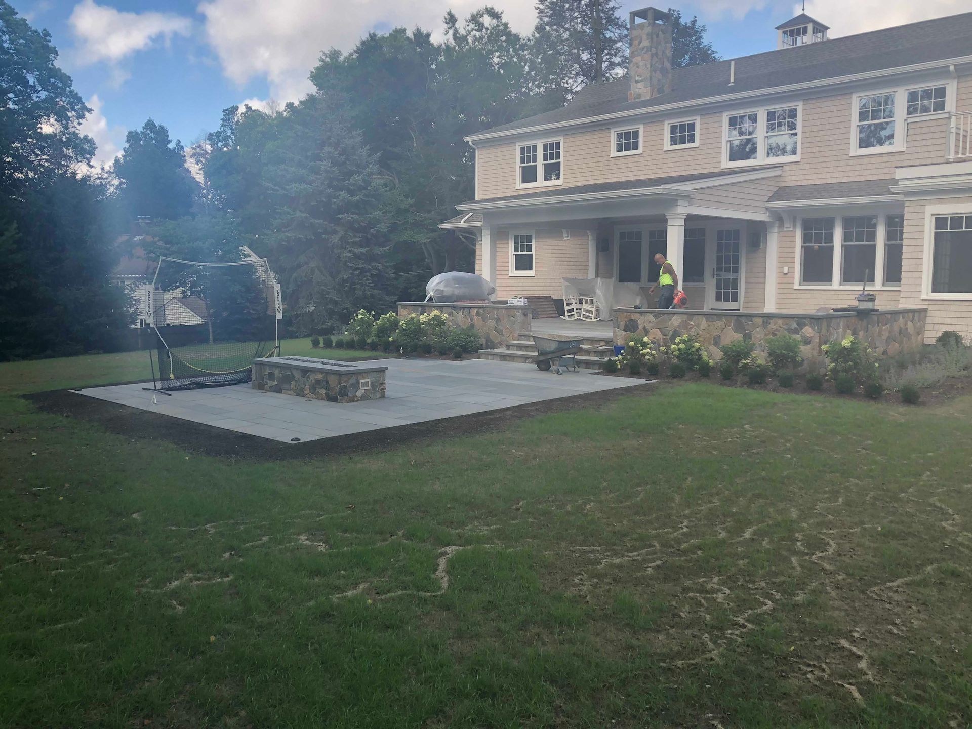 A worker in a yellow vest on a stone patio behind a large tan house with a green lawn in the foreground.