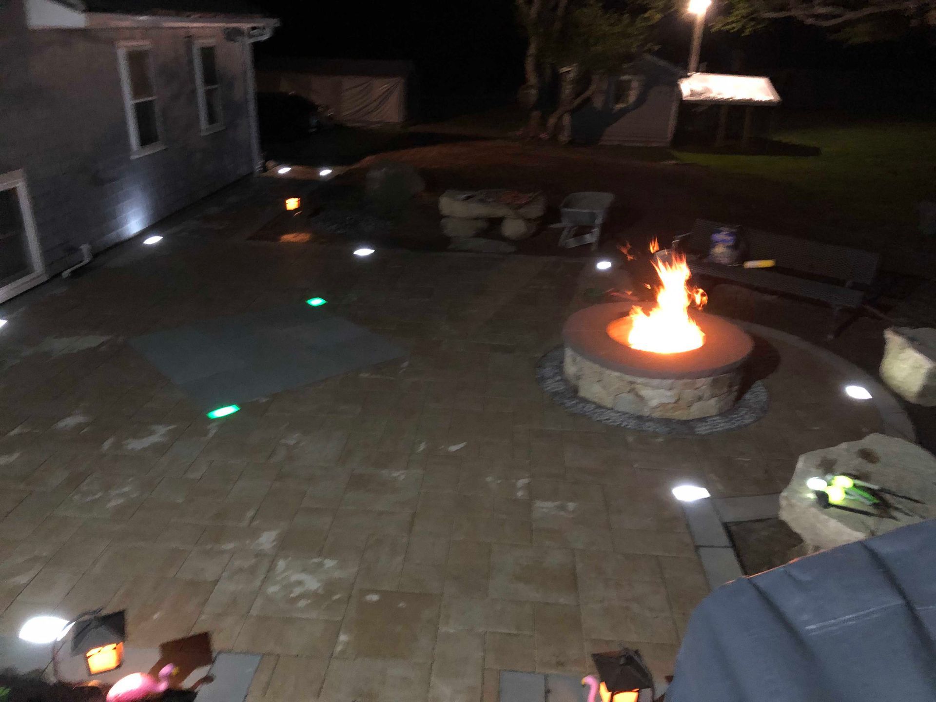 A fire pit burns in a stone patio at night, illuminated by soft in-ground lights and nearby outdoor lamps.