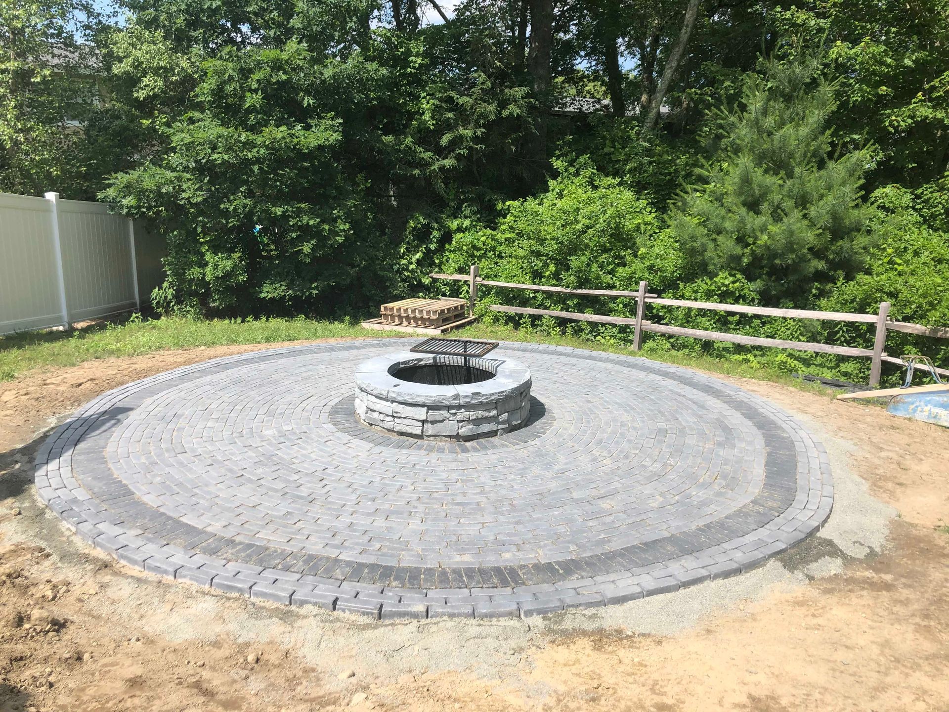 A circular paver patio with a central stone fire pit, set in a backyard near a wooden fence and trees.