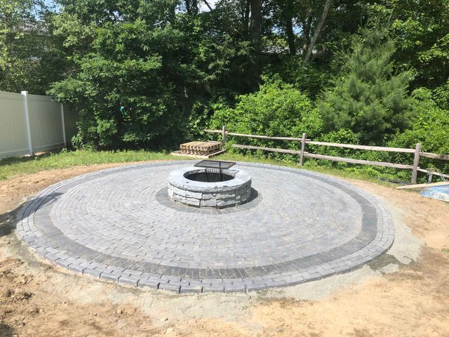 A circular stone paver patio with a central fire pit sits in a grassy backyard, surrounded by a split-rail fence.
