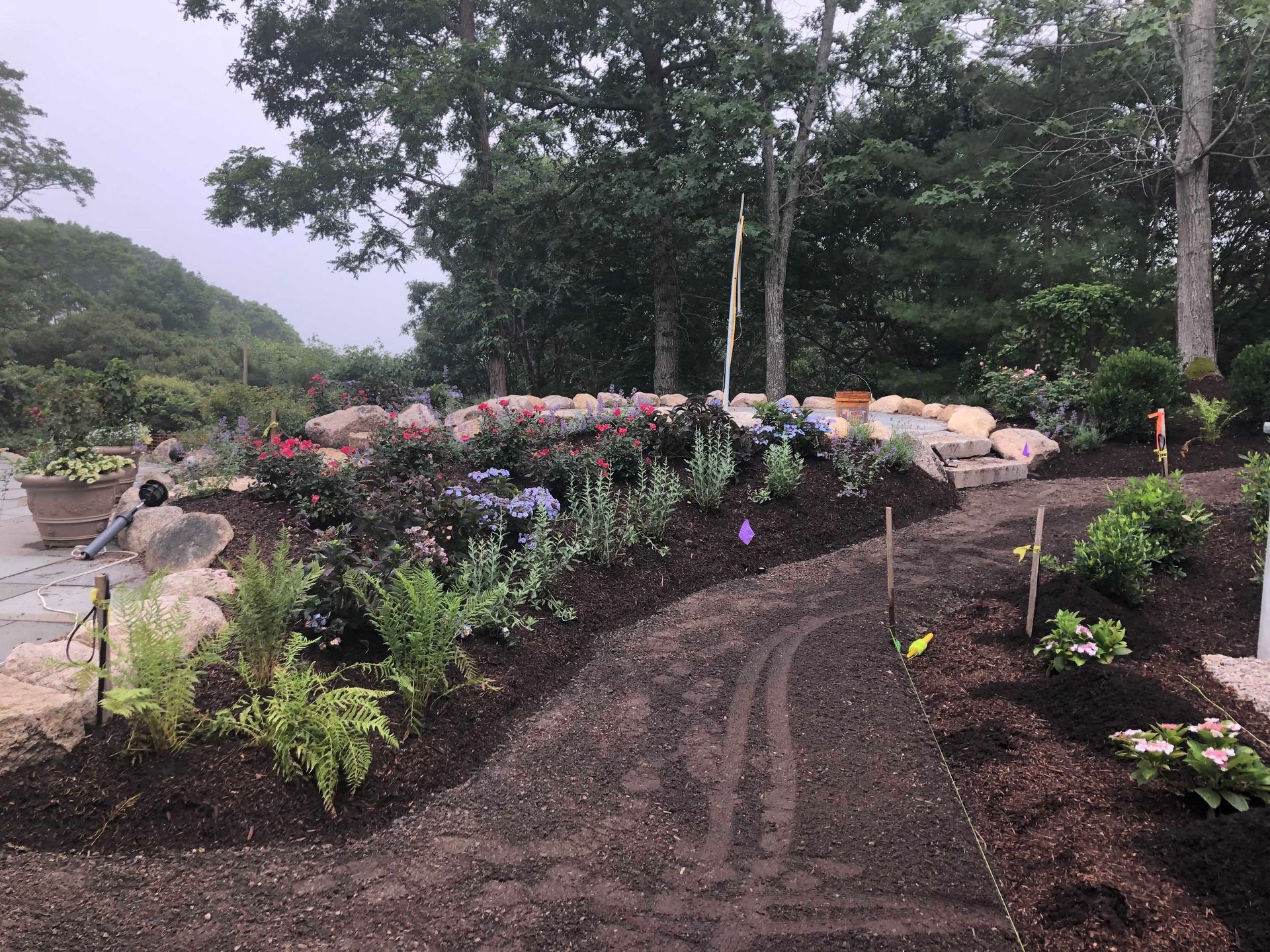 A dirt path curves through a garden featuring new plantings, dark mulch, and a tiered rock retaining wall under trees.