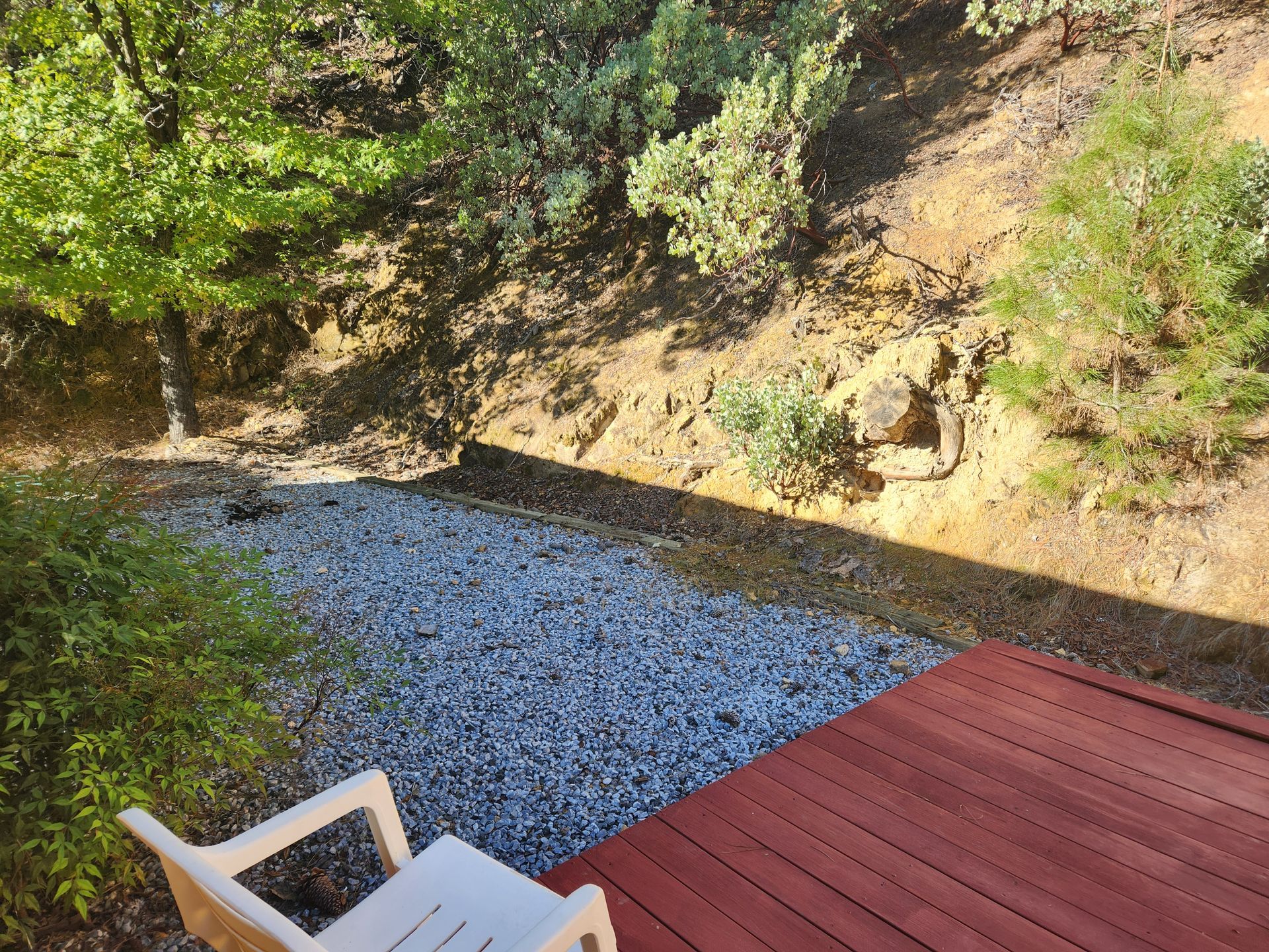 View from red deck onto gravel area and sloped, dirt hillside with trees. White plastic chair in foreground.