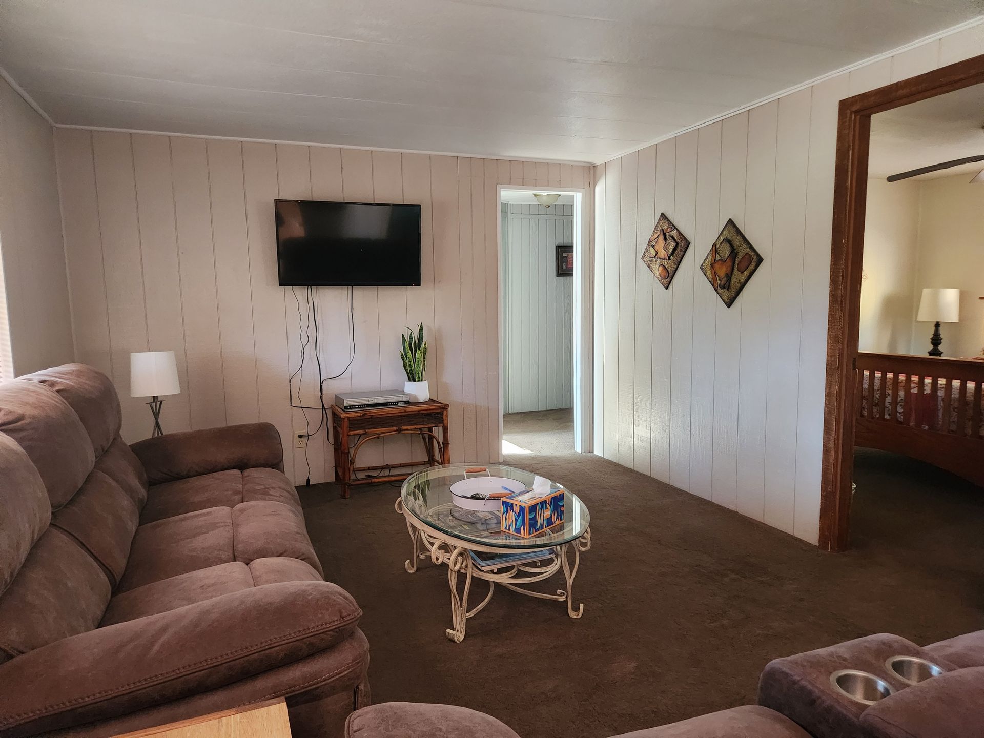 Living room with a brown recliner, TV, and coffee table. White paneled walls, and a doorway to another room.