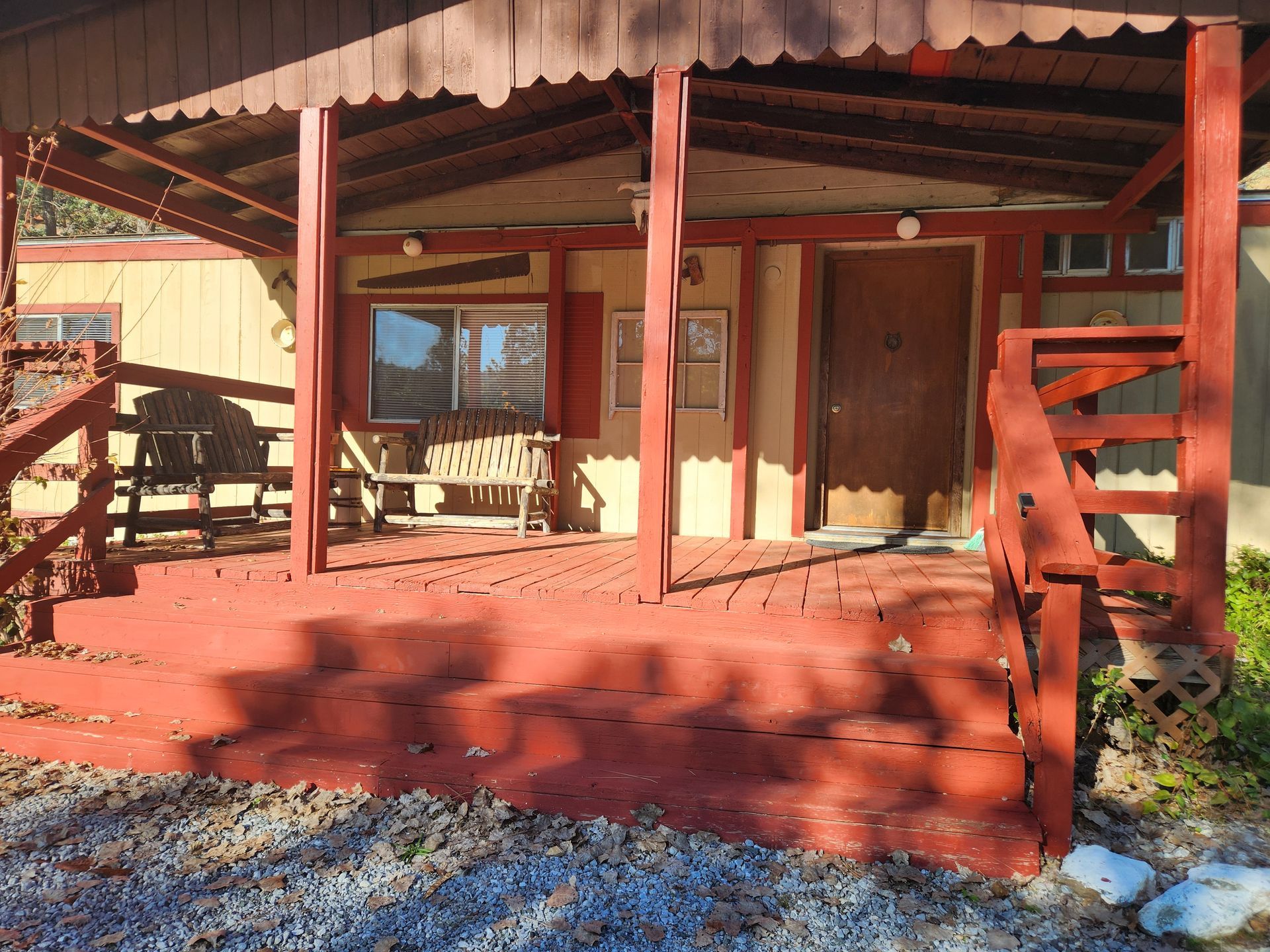 Red-painted porch of a tan house. Bench, window, and door under the roof. Gravel in foreground.