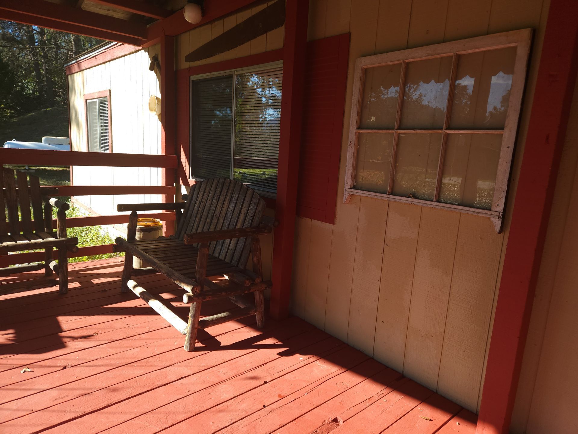 Wooden porch with a bench and a window. Red painted wood, sunlight, and a small building in the background.