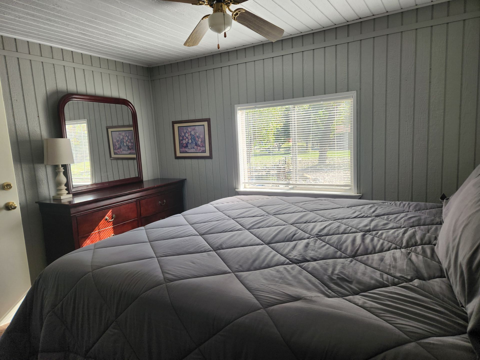 Bedroom with gray walls, bed, dresser, mirror, window. Sunlight streams through.