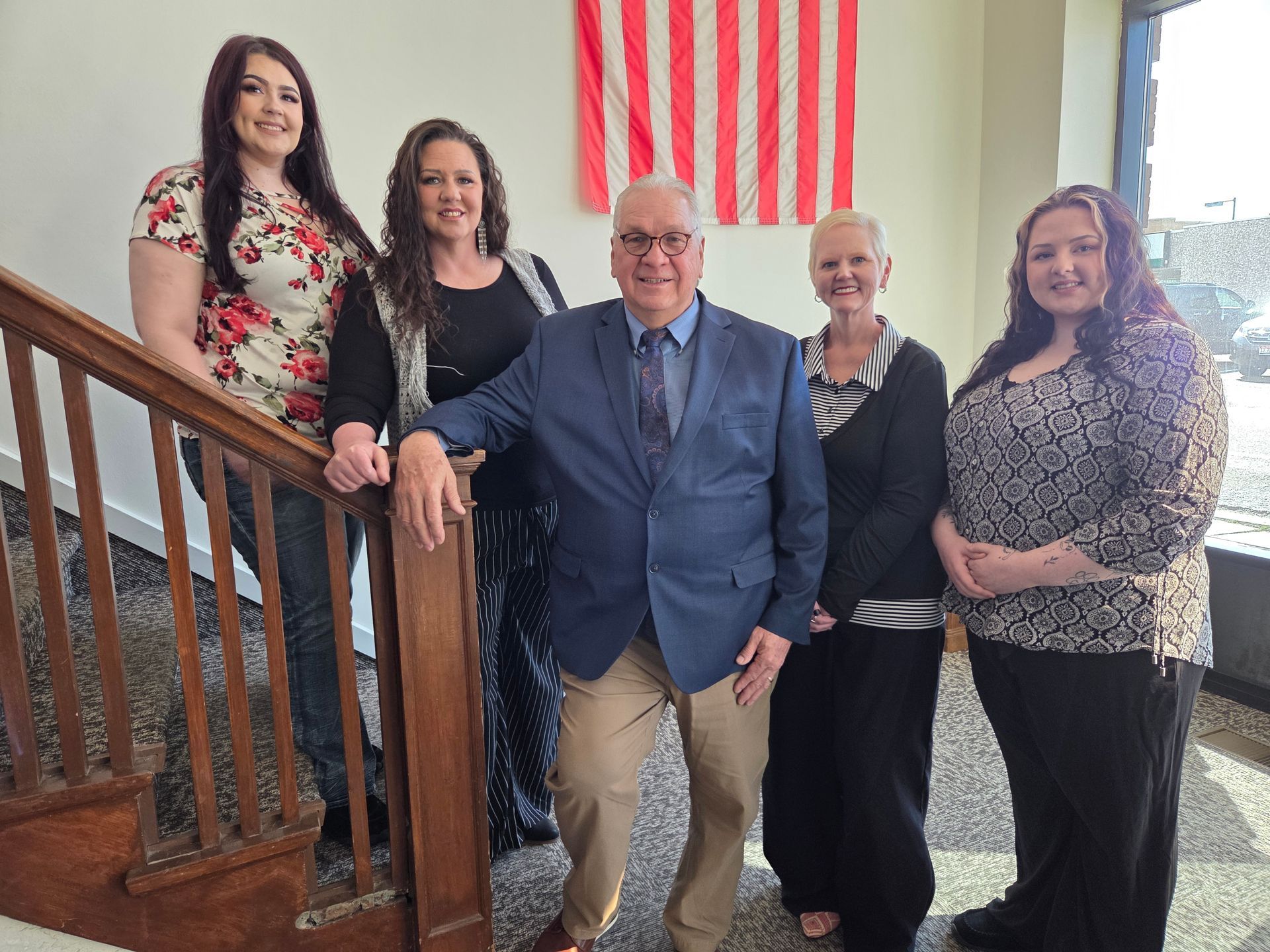 Five people standing indoors in front of an American flag, with a wooden staircase on the left side of the frame.